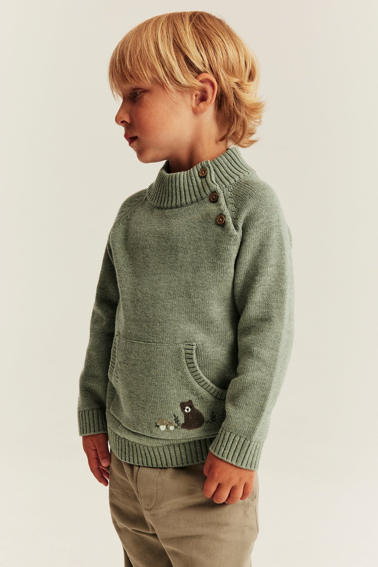 Seitenansicht eines Kindes in grünem Strickpullover mit Knöpfen am Kragen und Bärenstickerei.