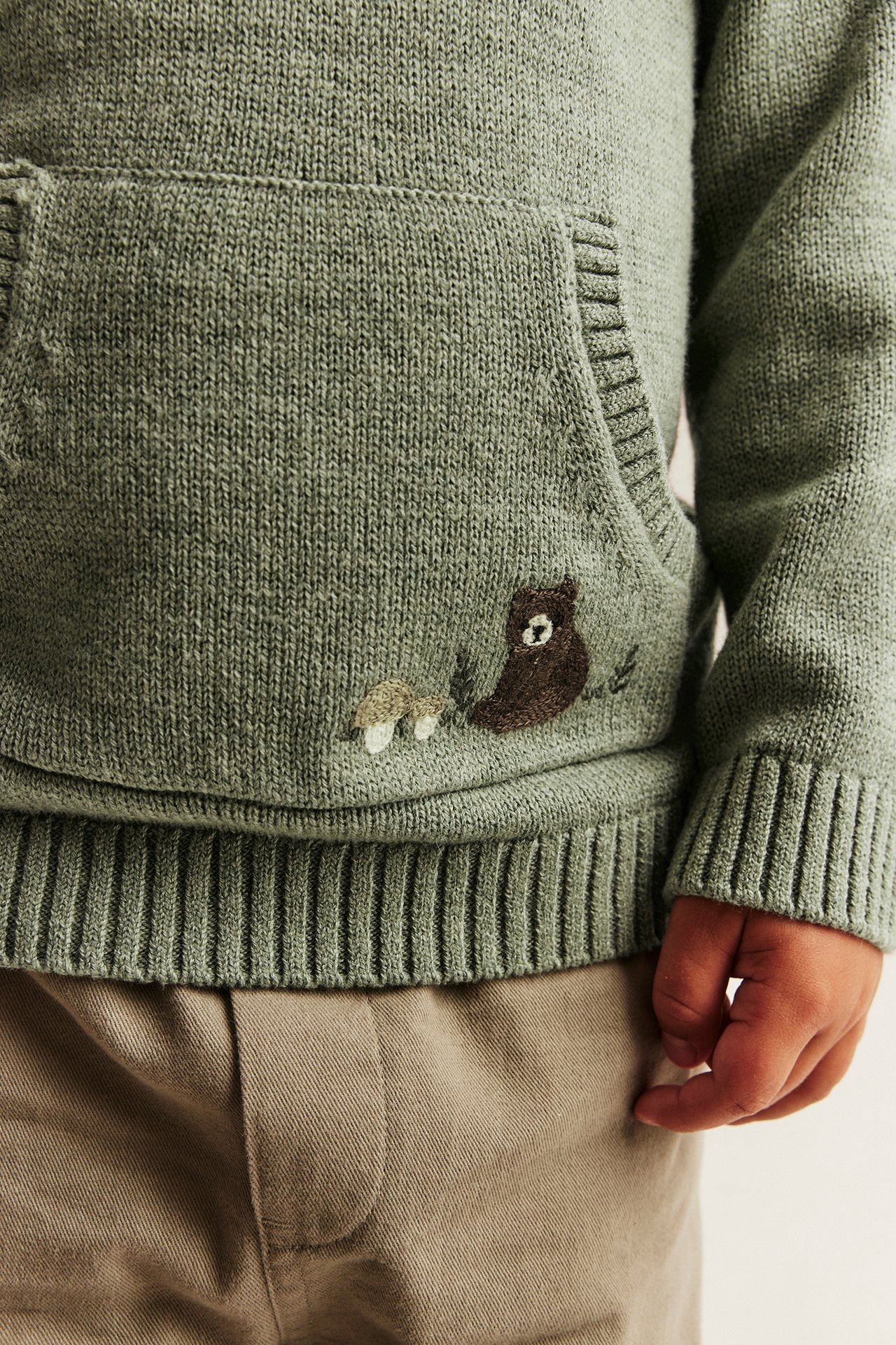 Nahaufnahme vorne von Kinder grüner Strick-Kapuzenpullover mit gesticktem Bären und Pilzen, und brauner Hose.
