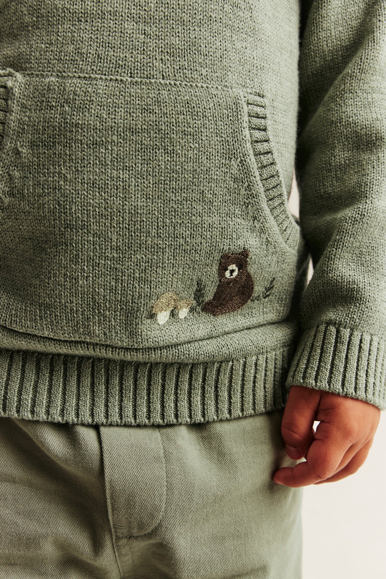 Nahaufnahme eines grünen Strick-Kapuzenpullovers für Kinder mit Bär- und Pilzstickerei und grüner Hose.