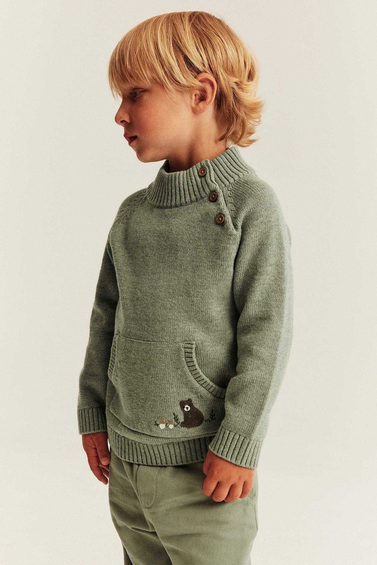 Seitenansicht: Grüner Kinderstrickpullover mit Knopfkragen, Kängurutasche und Bärenstickerei.