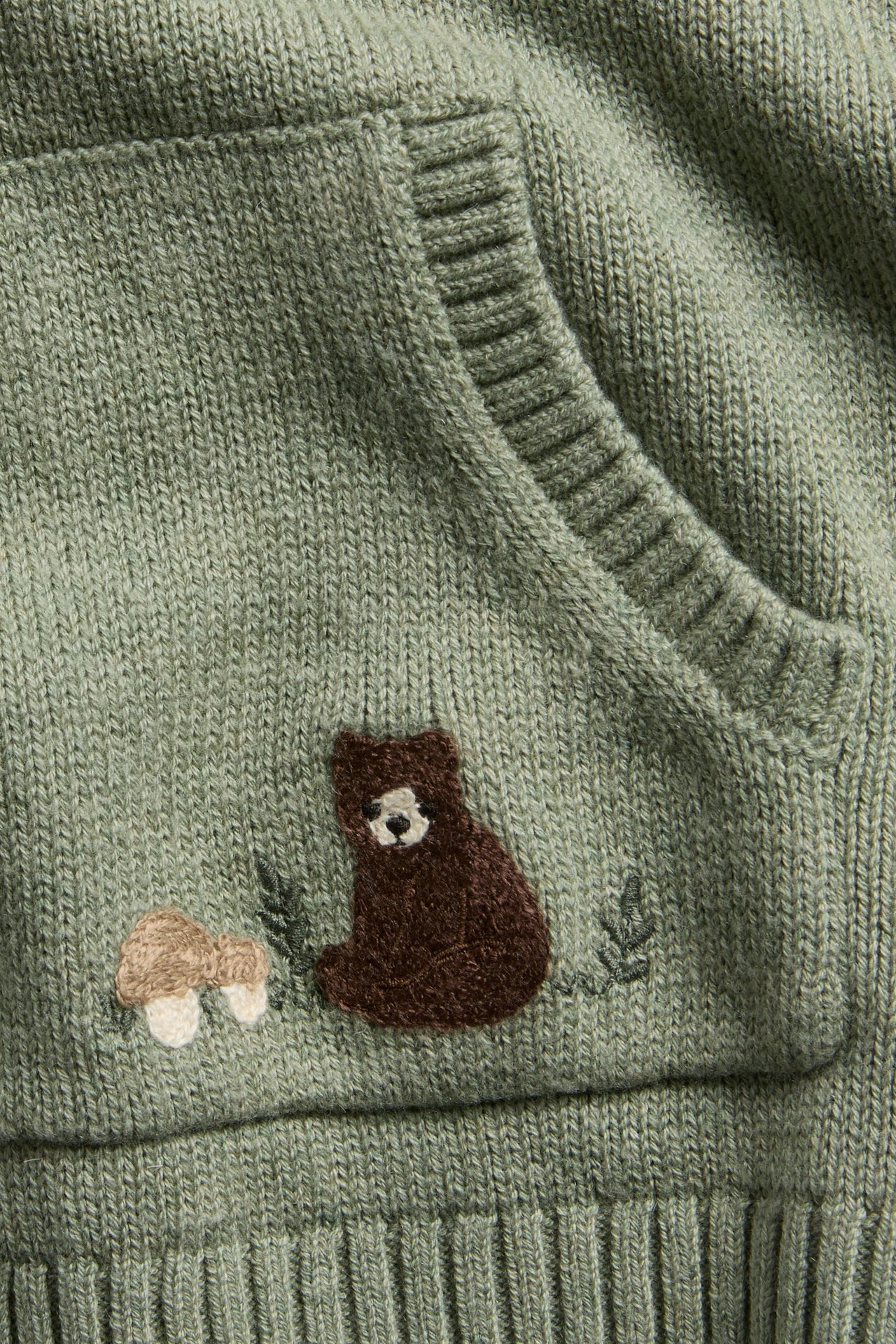 Nahaufnahme einer grün gestrickten Kinderkleidertasche mit gesticktem braunem Bären und Pilzen.