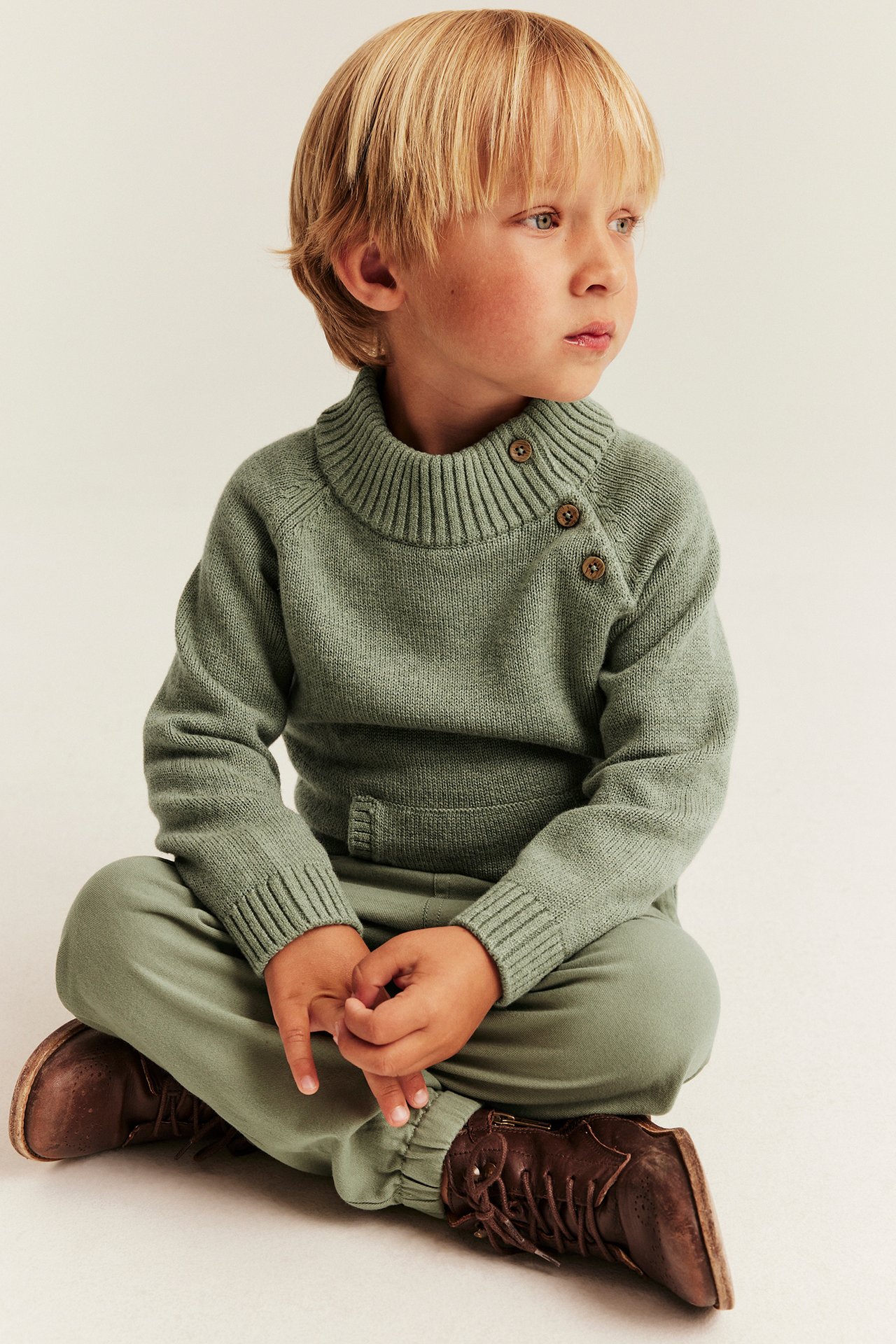 Vorderansicht: Kind in salbeigrünem Strickpullover mit Rollkragen und Knöpfen, salbeigrüner Jogginghose und braunen Stiefeln.