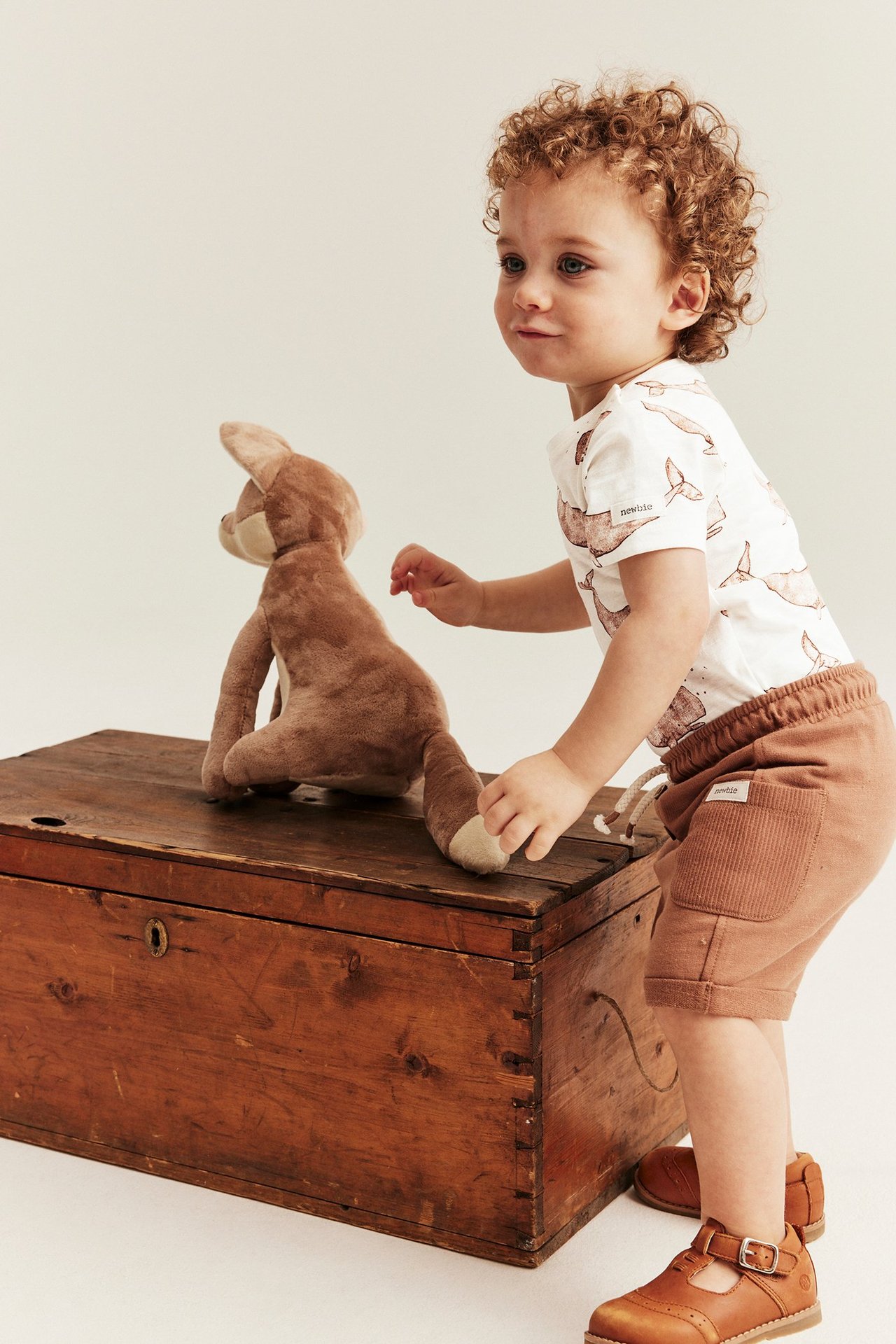 Baby in cremefarbenem kurzärmeligem Body mit Walmuster, braunen Shorts und braunen Schnallenschuhen.