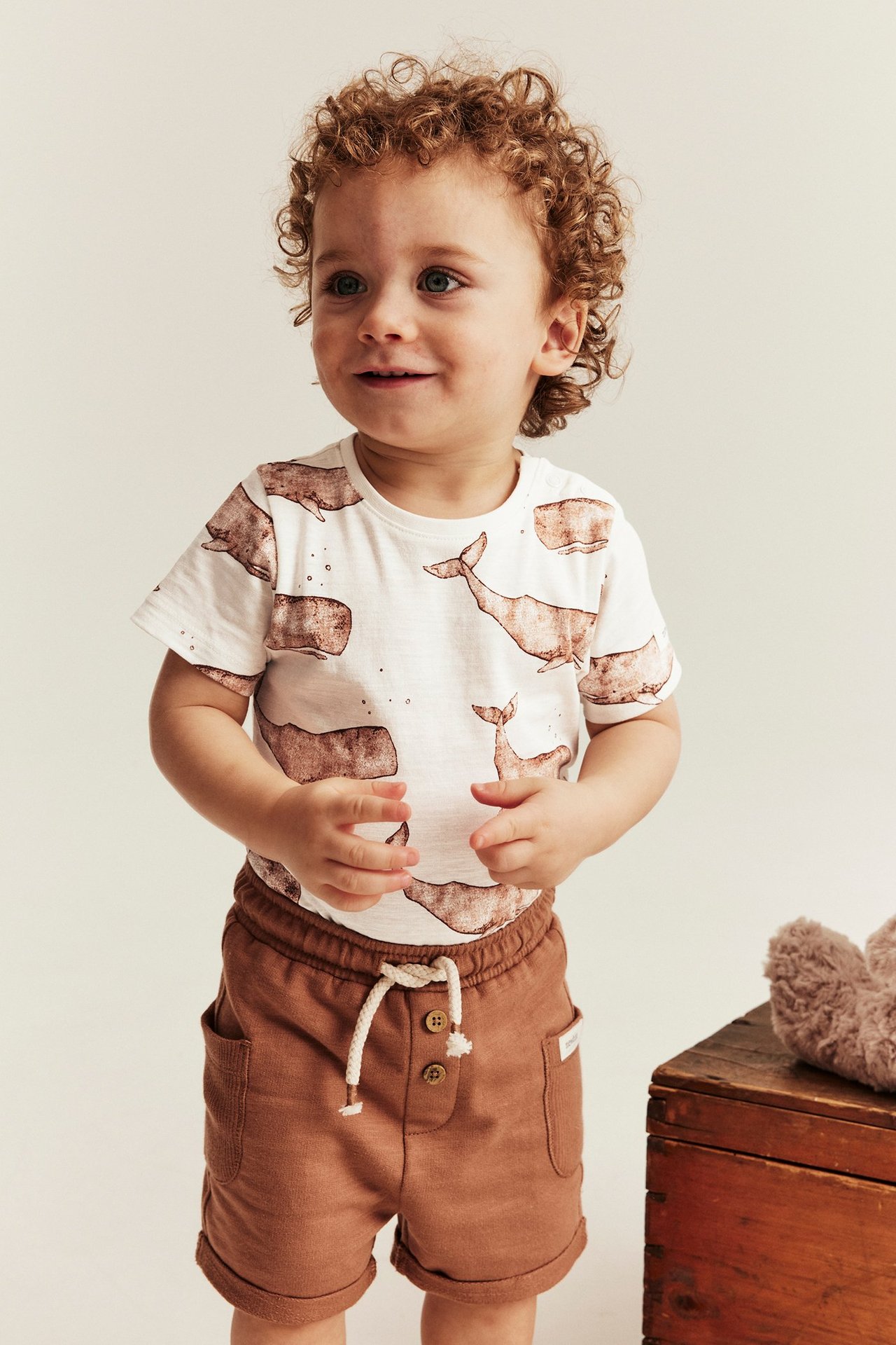 Vorderansicht: Baby in cremefarbenem kurzärmeligem Baumwoll-Body mit Walmuster und braunen Shorts.