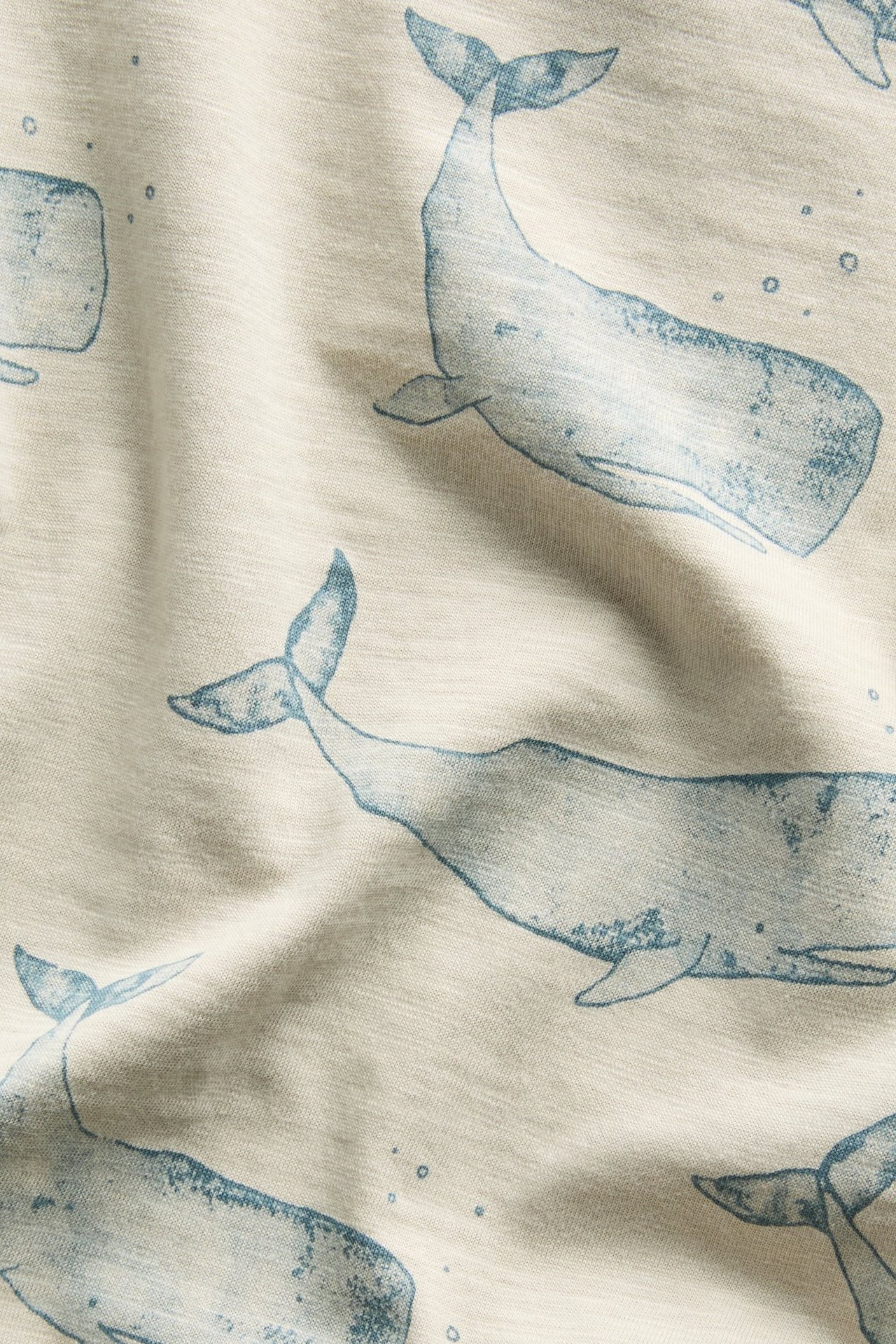 Nahaufnahme eines beigen Baumwoll-Slub-Jersey Babybodys mit blauem Wal- und Blasenmuster.