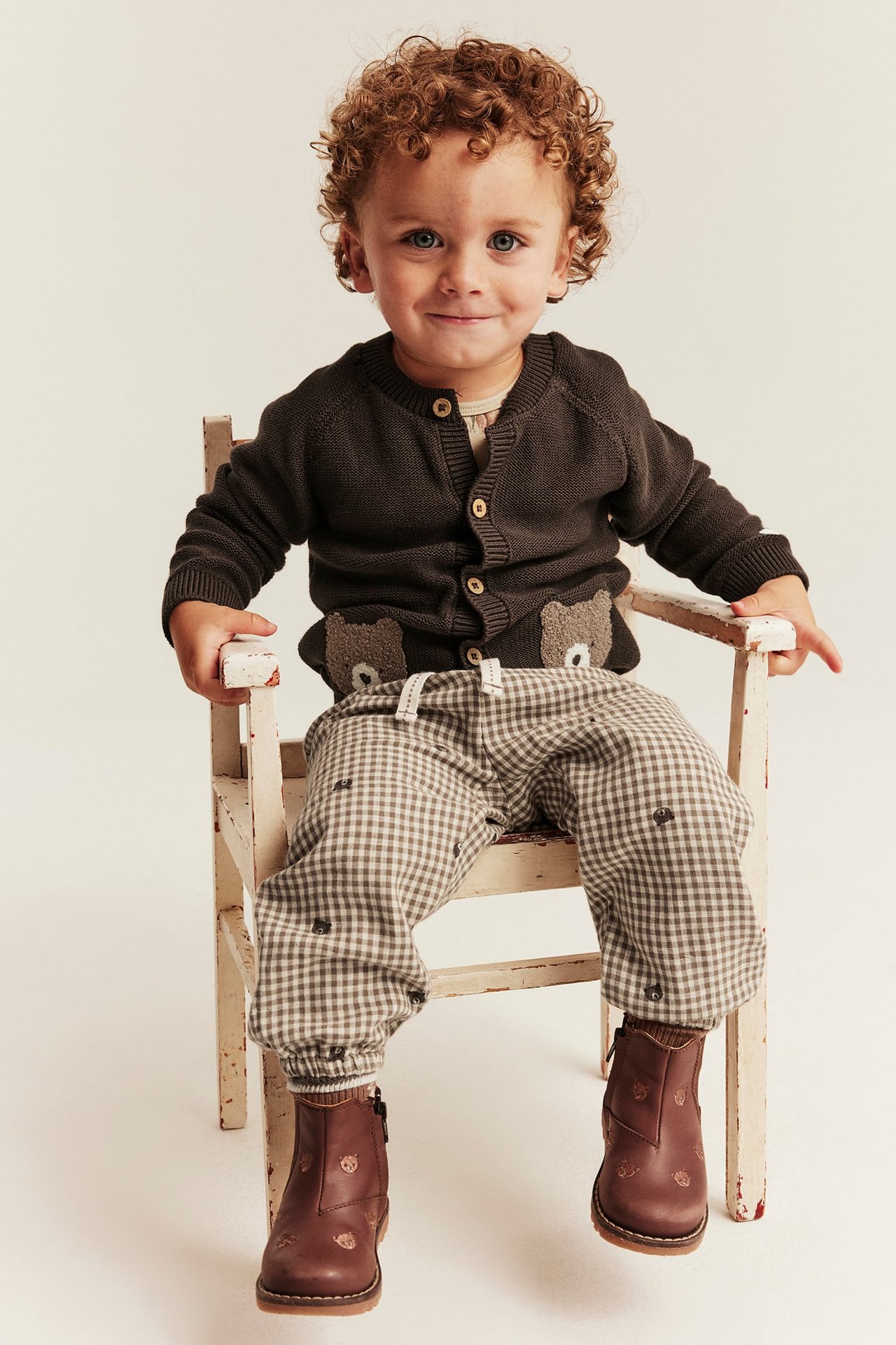Front view of a kid wearing a dark brown knit button cardigan with bear pockets, brown plaid pants, and brown leather ankle boots.