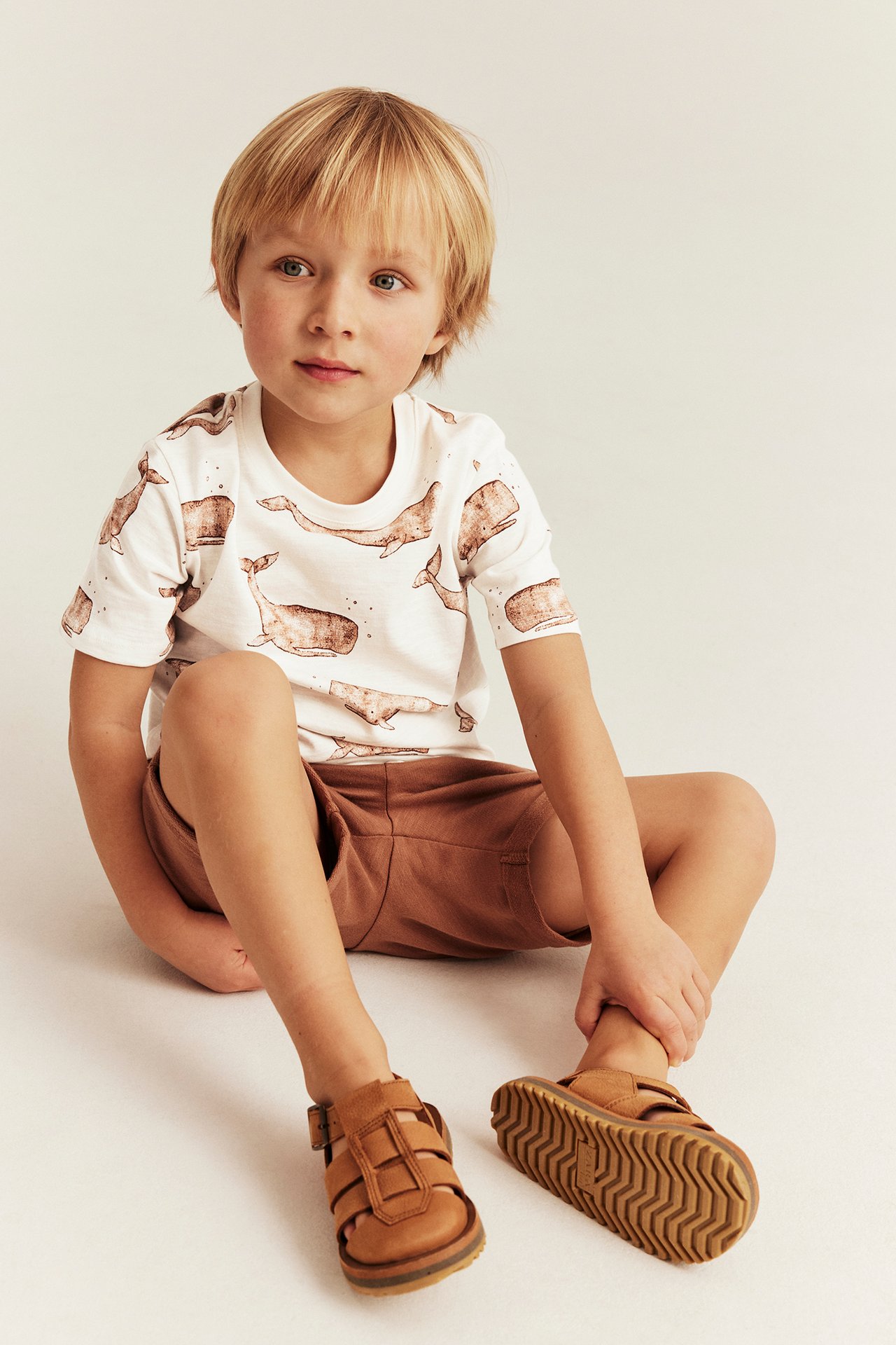 Vorderansicht eines Kindes mit einem cremeweißen Baumwoll-T-Shirt mit Walmuster, braunen Shorts und braunen Sandalen.