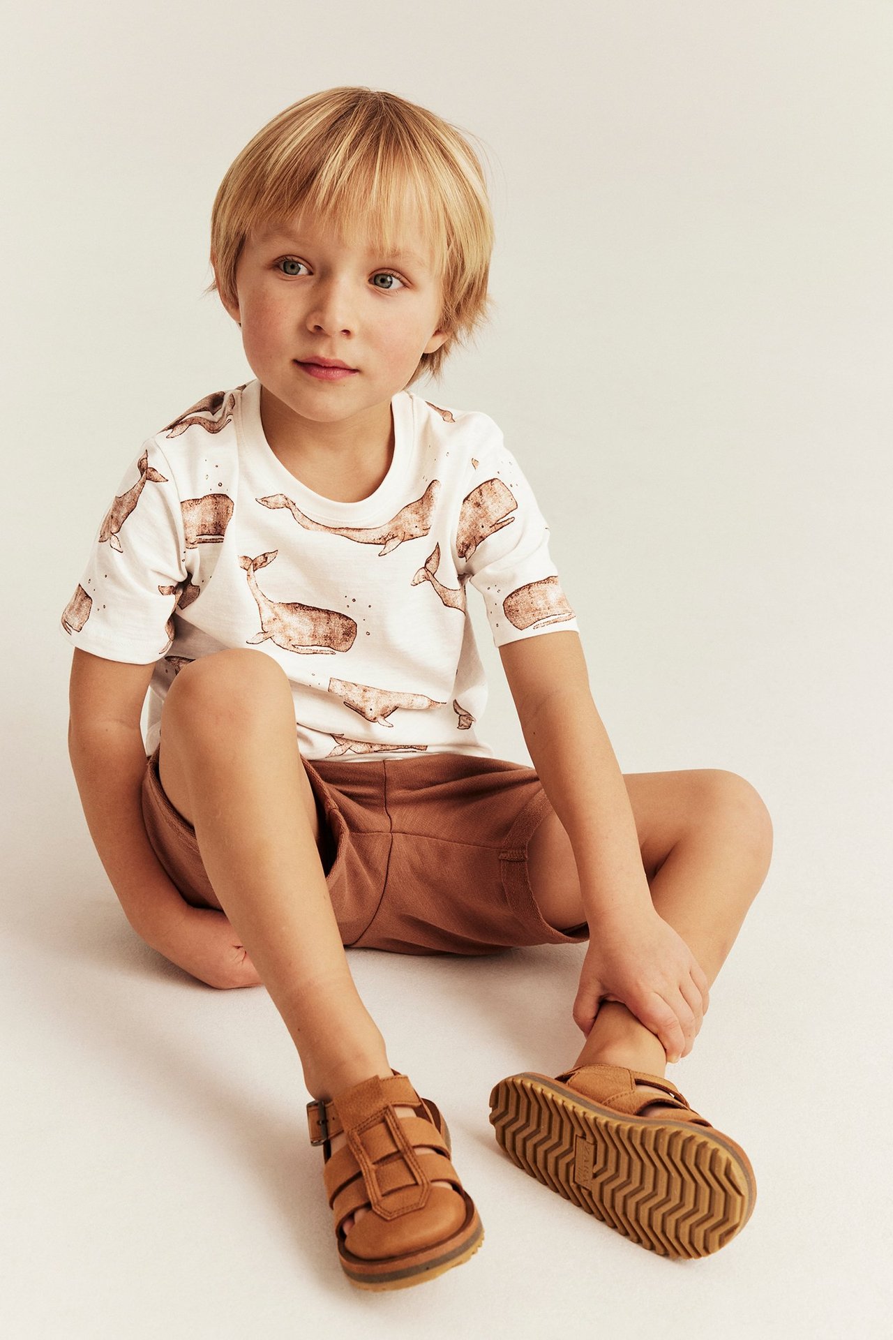 Vorderansicht eines Kindes mit einem cremeweißen Baumwoll-T-Shirt mit Walmuster, braunen Shorts und braunen Sandalen.