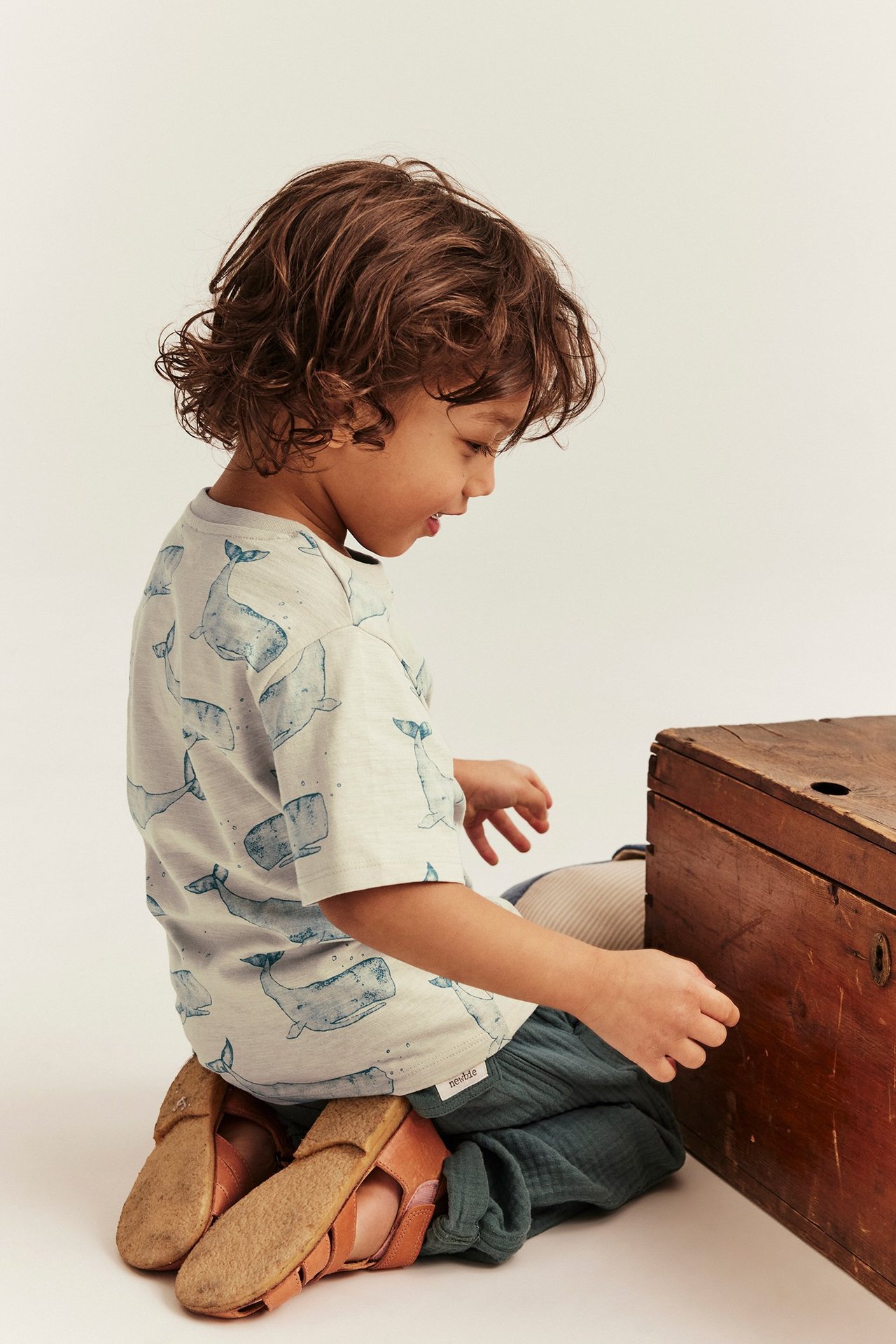 Seitenansicht eines Kindes in beigem Slub-T-Shirt mit Walmuster, dunkelgrüner Hose und braunen Sandalen.