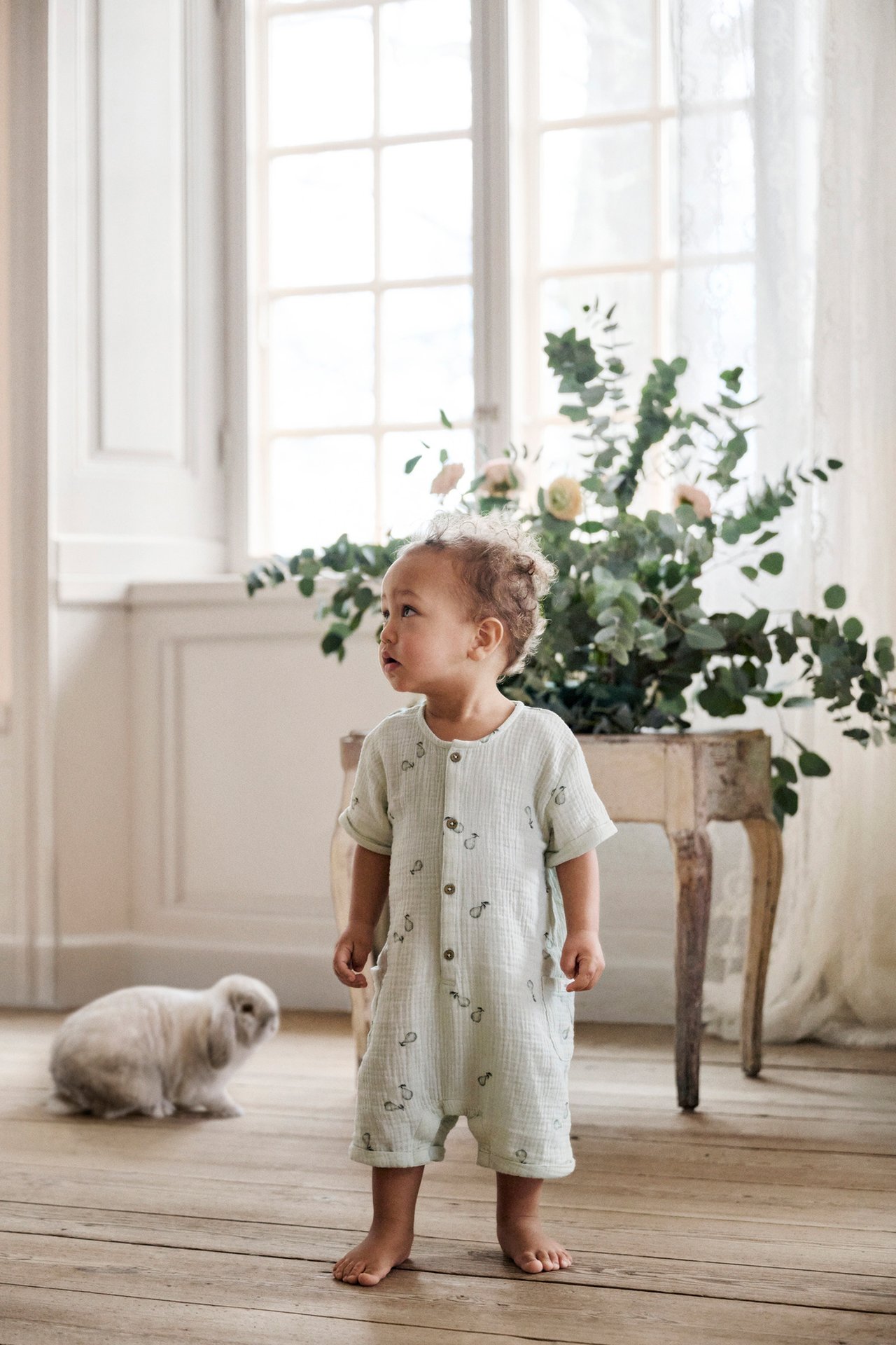 Front view of a baby wearing a light green cotton pear-patterned romper with front buttons and patch pockets.