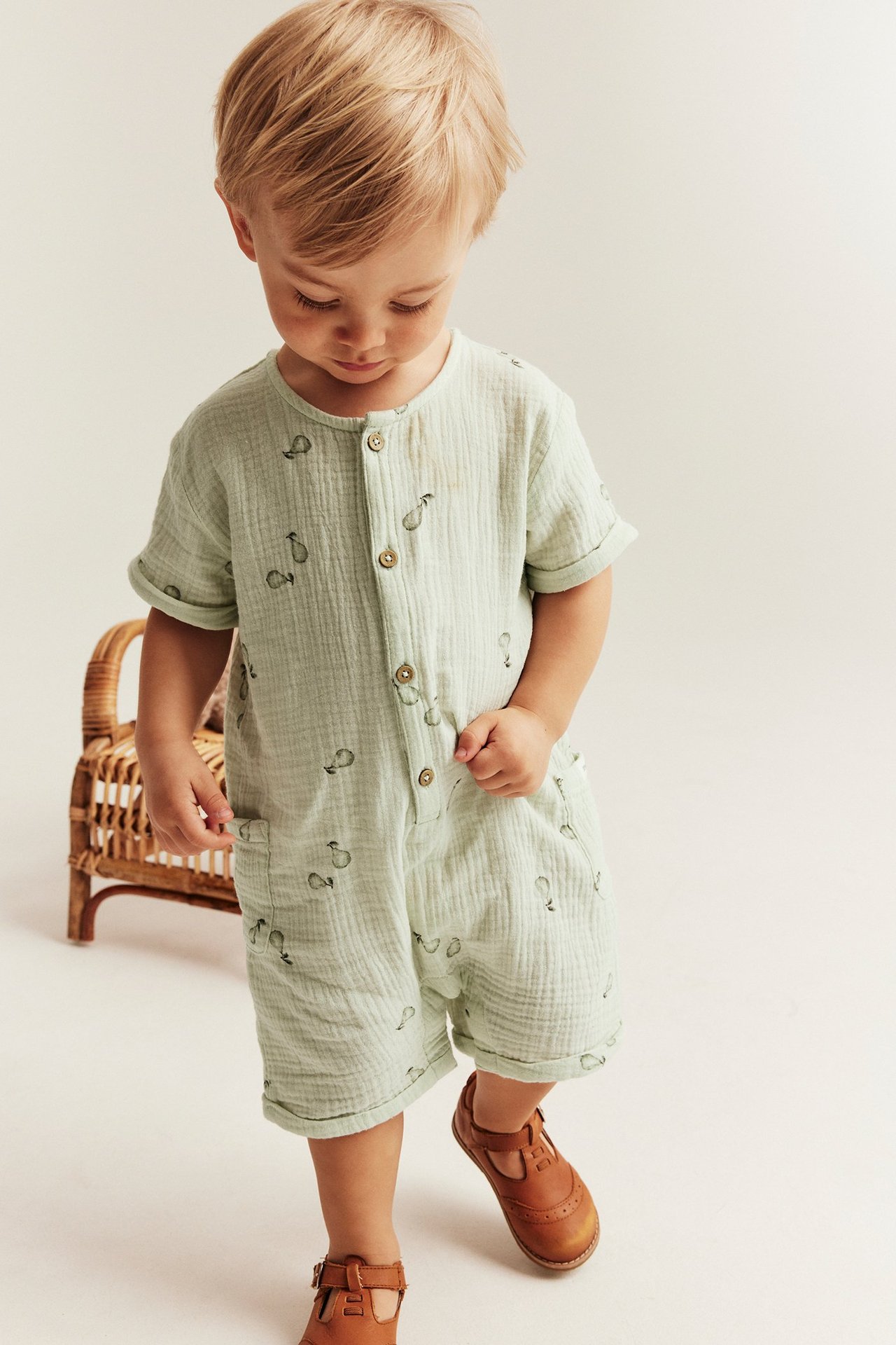 Vorderansicht, Baby trägt hellgrünen Birnenmuster-Baumwoll-Romper mit Knöpfen, Taschen und braunen Schuhen.