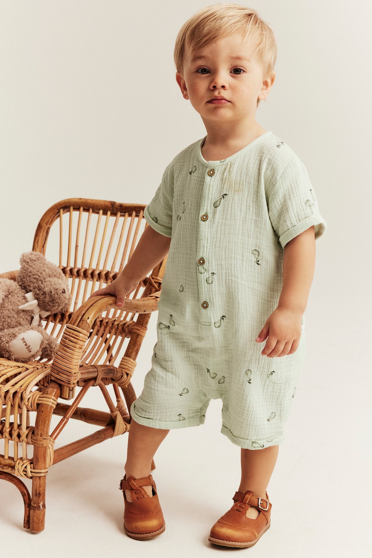Vorderansicht: Kind in hellgrünem Baumwoll-Strampler mit Birnenmuster, Knöpfen und braunen T-Steg-Schuhen.