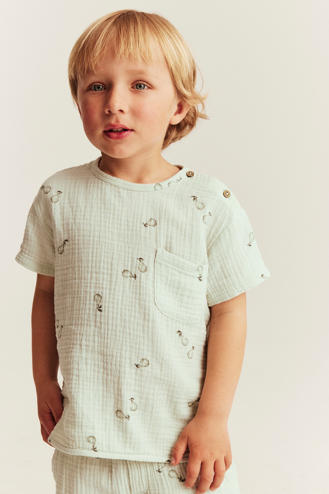 Kid wears a light green pear-patterned muslin top with shoulder buttons and chest pocket.