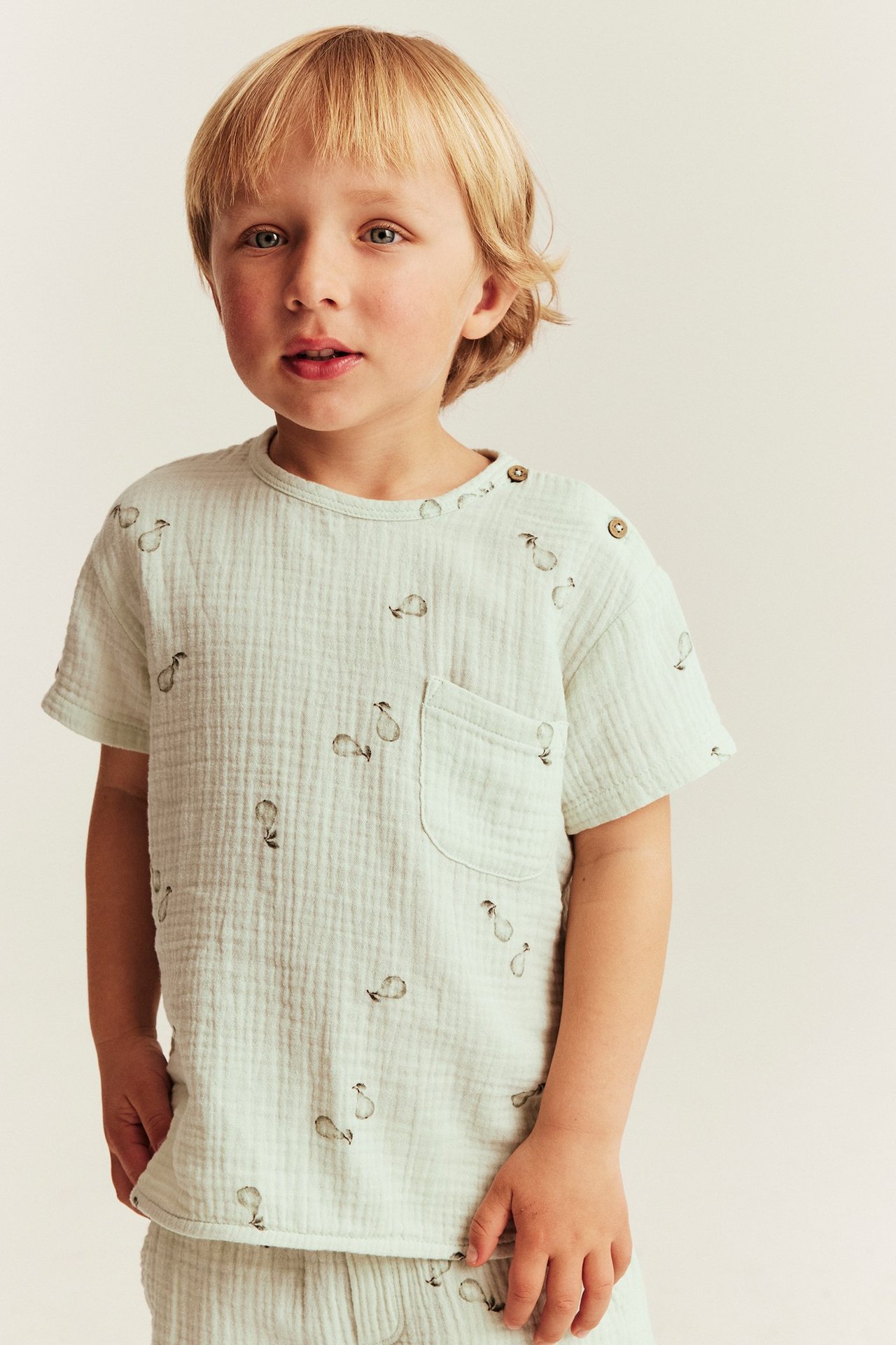 Kind trägt ein hellgrünes birnenmusteriges Musselin-Oberteil mit Schulterknöpfen und Brusttasche.