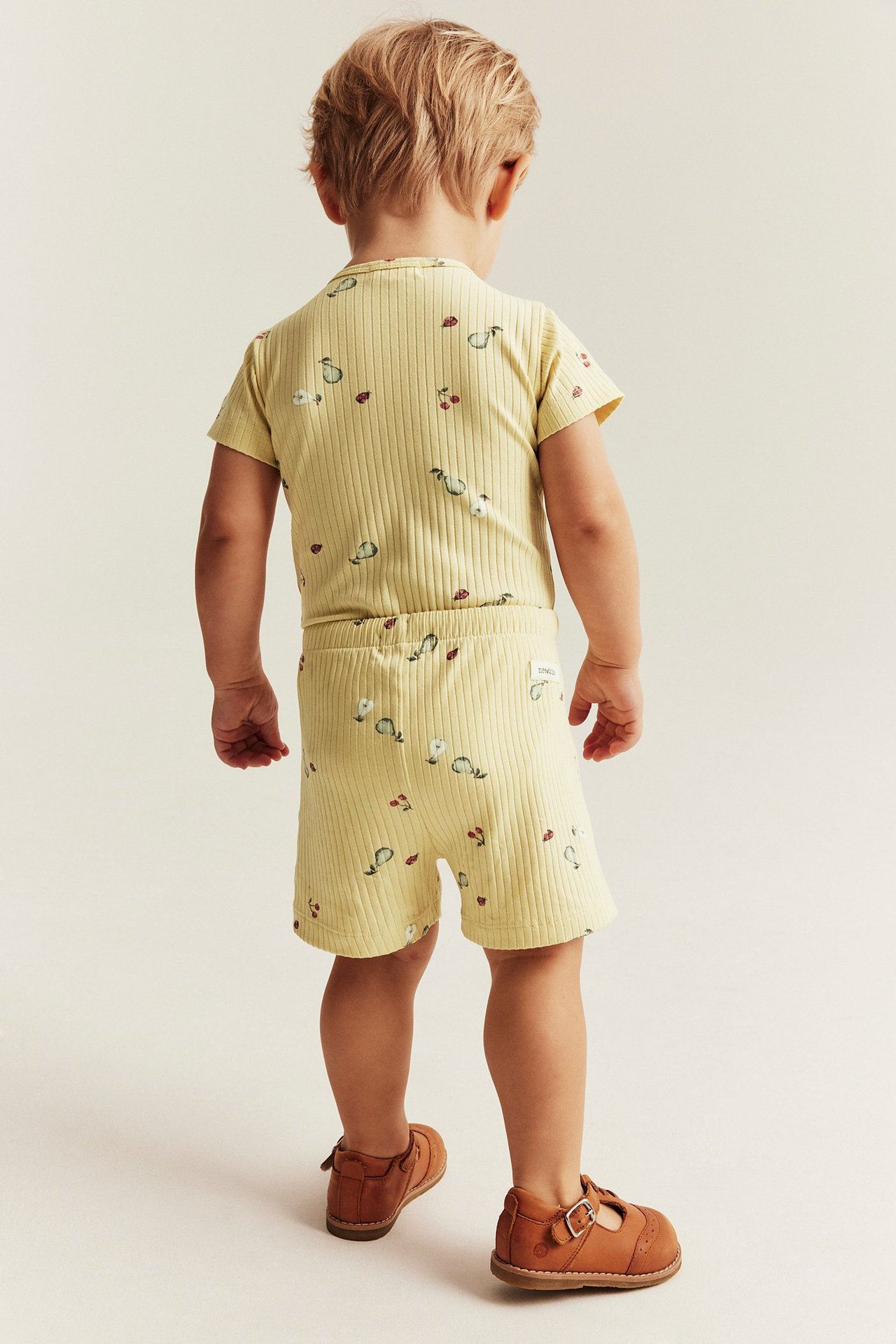 Rückenansicht: Kind in gelbem geripptem 2-teiligen Set mit Fruchtmuster und braunen Schnallenschuhen.