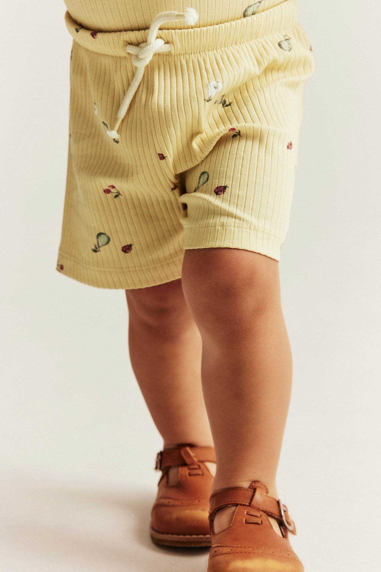 Close-up of baby wearing yellow ribbed shorts with fruit and ladybug print, white drawstring, and brown buckled shoes.
