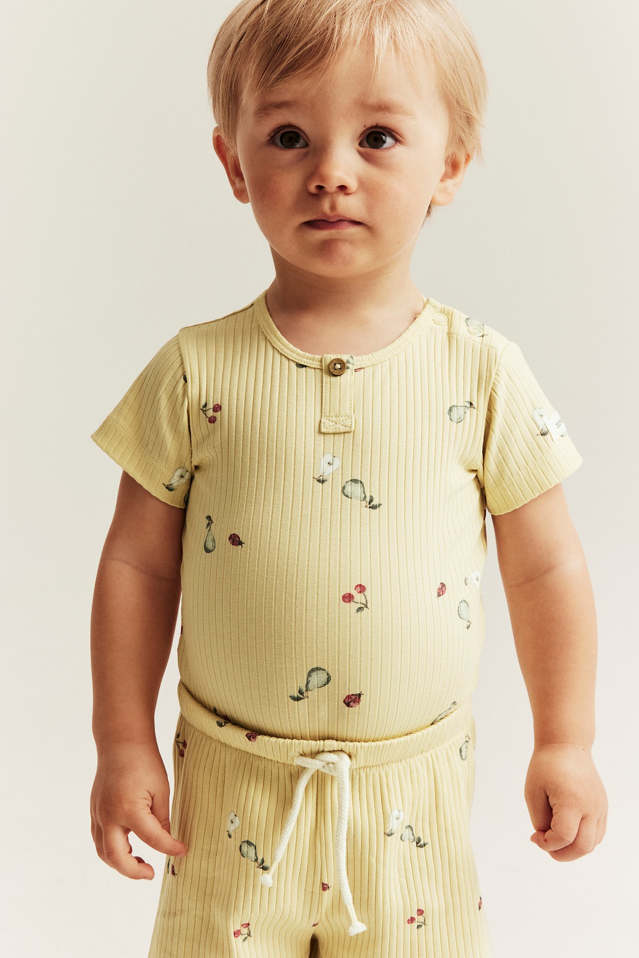 Front view: Baby wears a yellow ribbed 2-piece set with pear, cherry, and ladybug print.