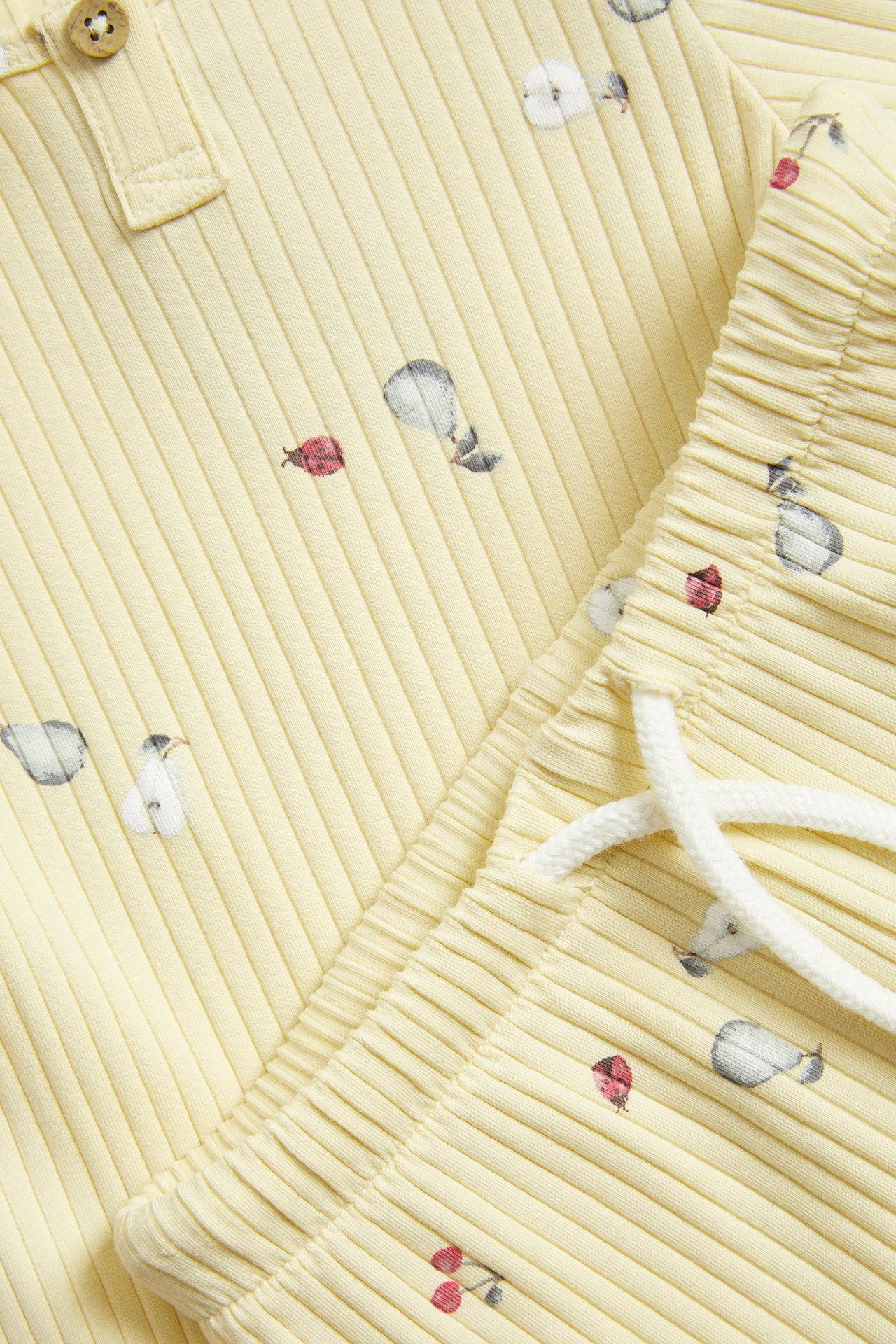 Close-up of a yellow ribbed two-piece set, patterned with pears, cherries, and ladybugs. Features buttons and a white drawstring.