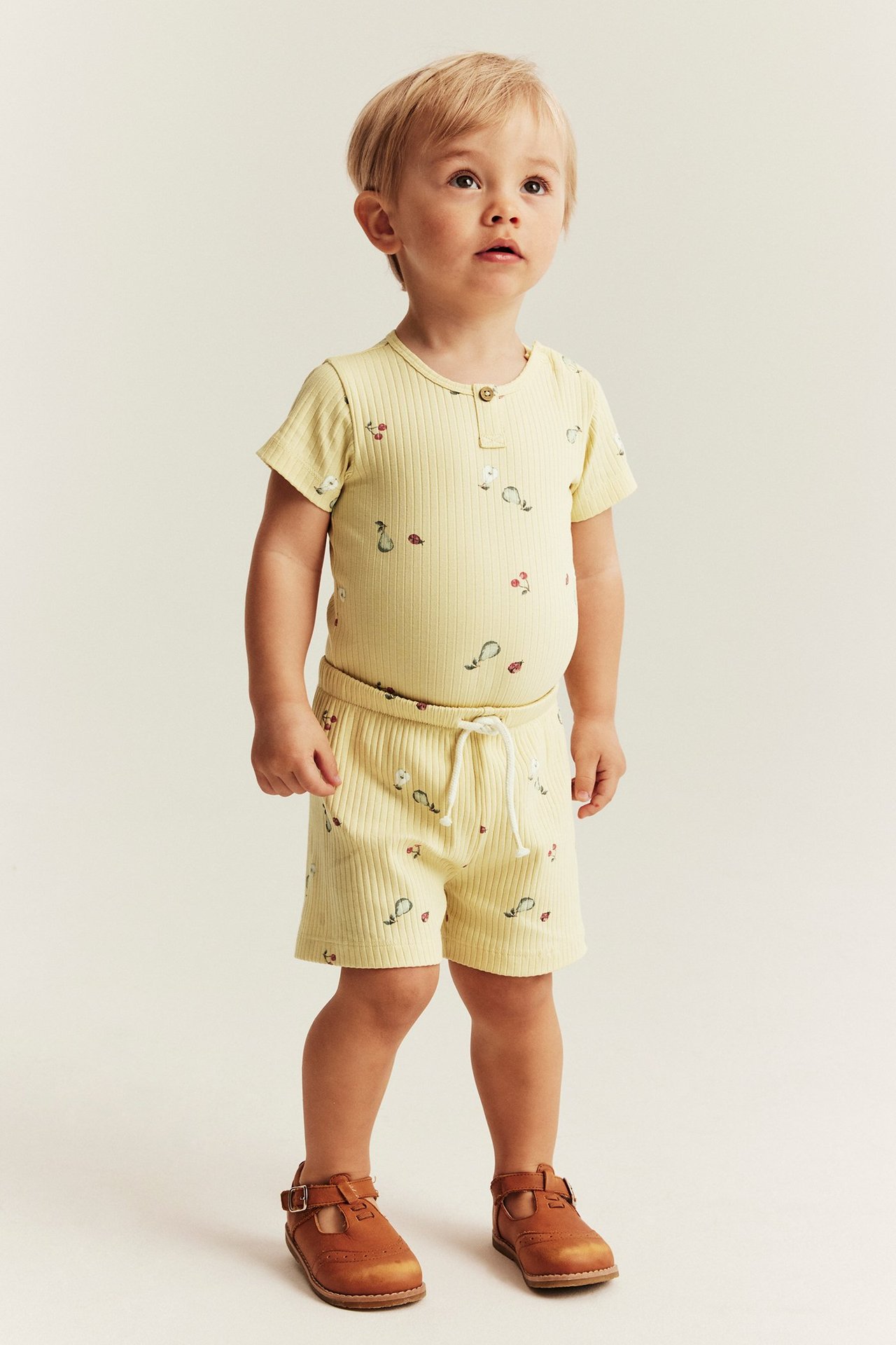 Frontansicht: Baby trägt gelben, gerippten Body und Shorts mit Obst- und Marienkäfer-Print.