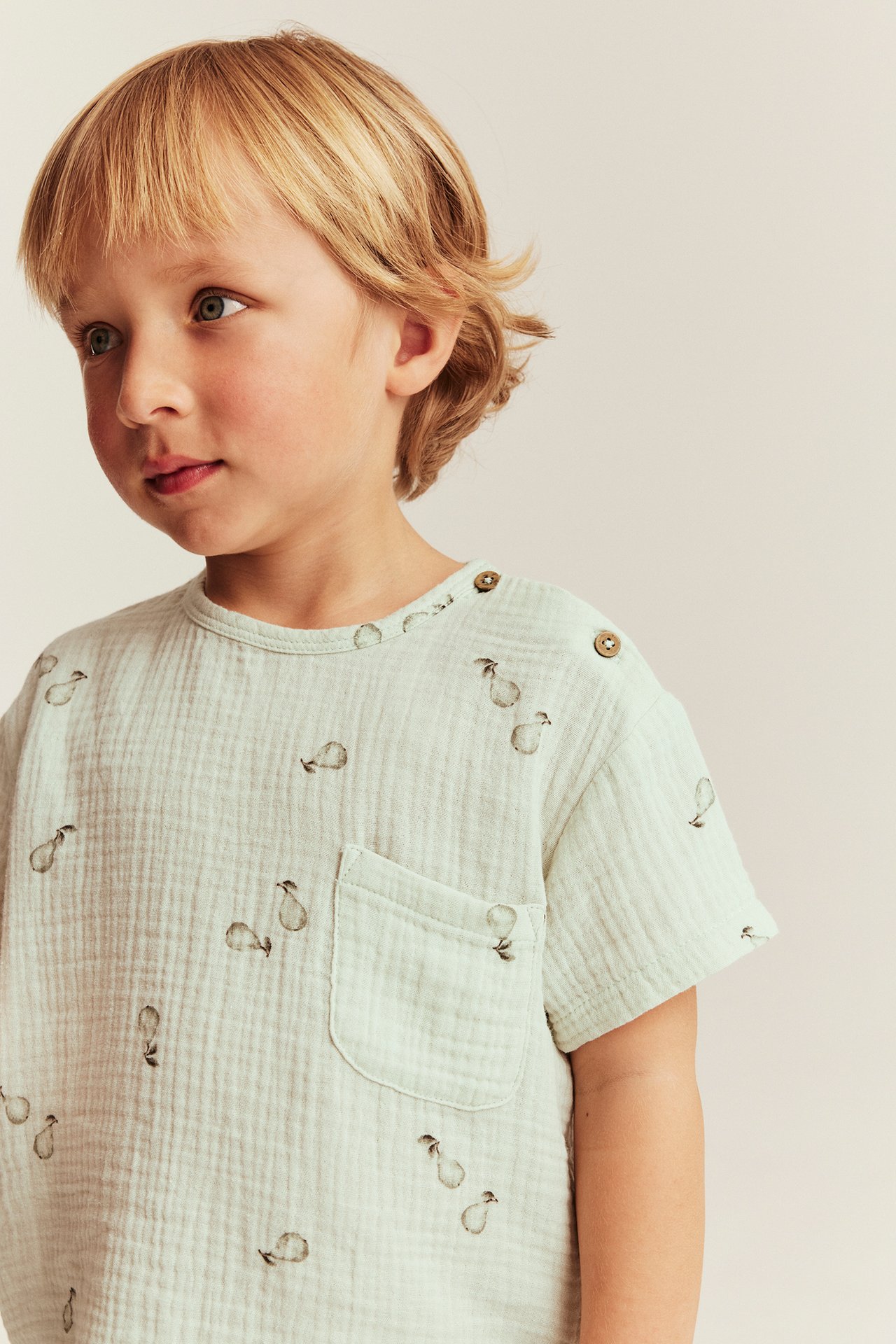 Front view: Kid in light green muslin t-shirt with pear pattern, wooden shoulder buttons, and chest pocket.