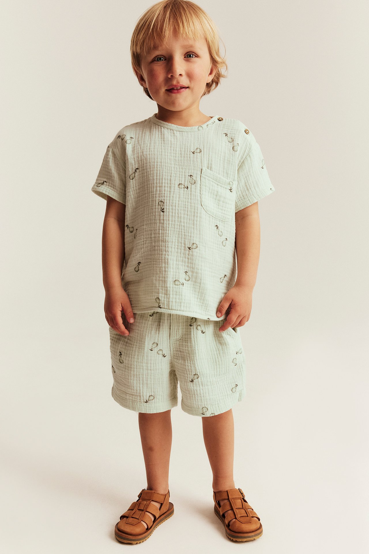 Front view of a kid wearing light green patterned muslin shorts, a matching t-shirt, and brown sandals.