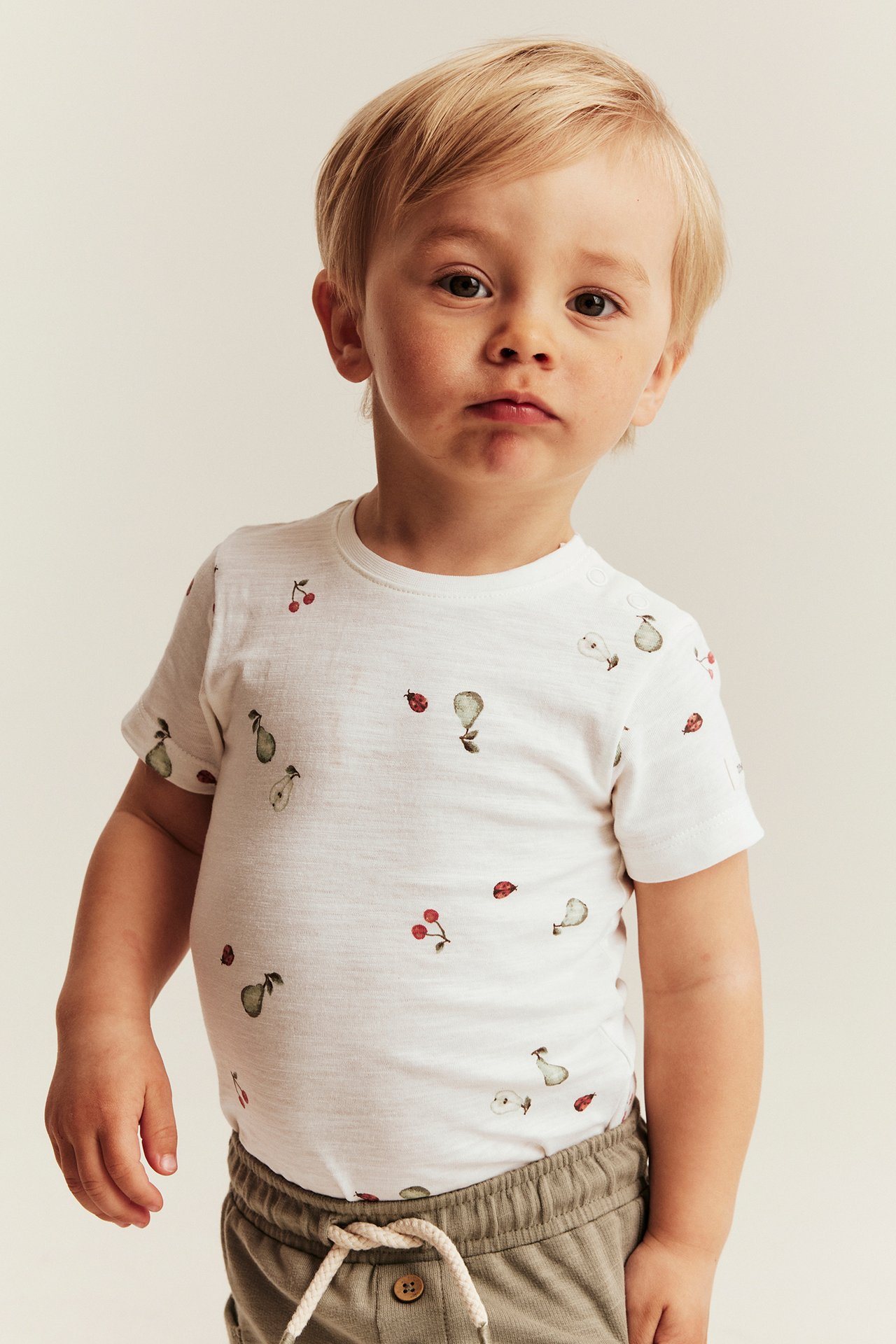 Front view of a kid wearing an off-white short-sleeved body with fruit and ladybug print, and khaki shorts.