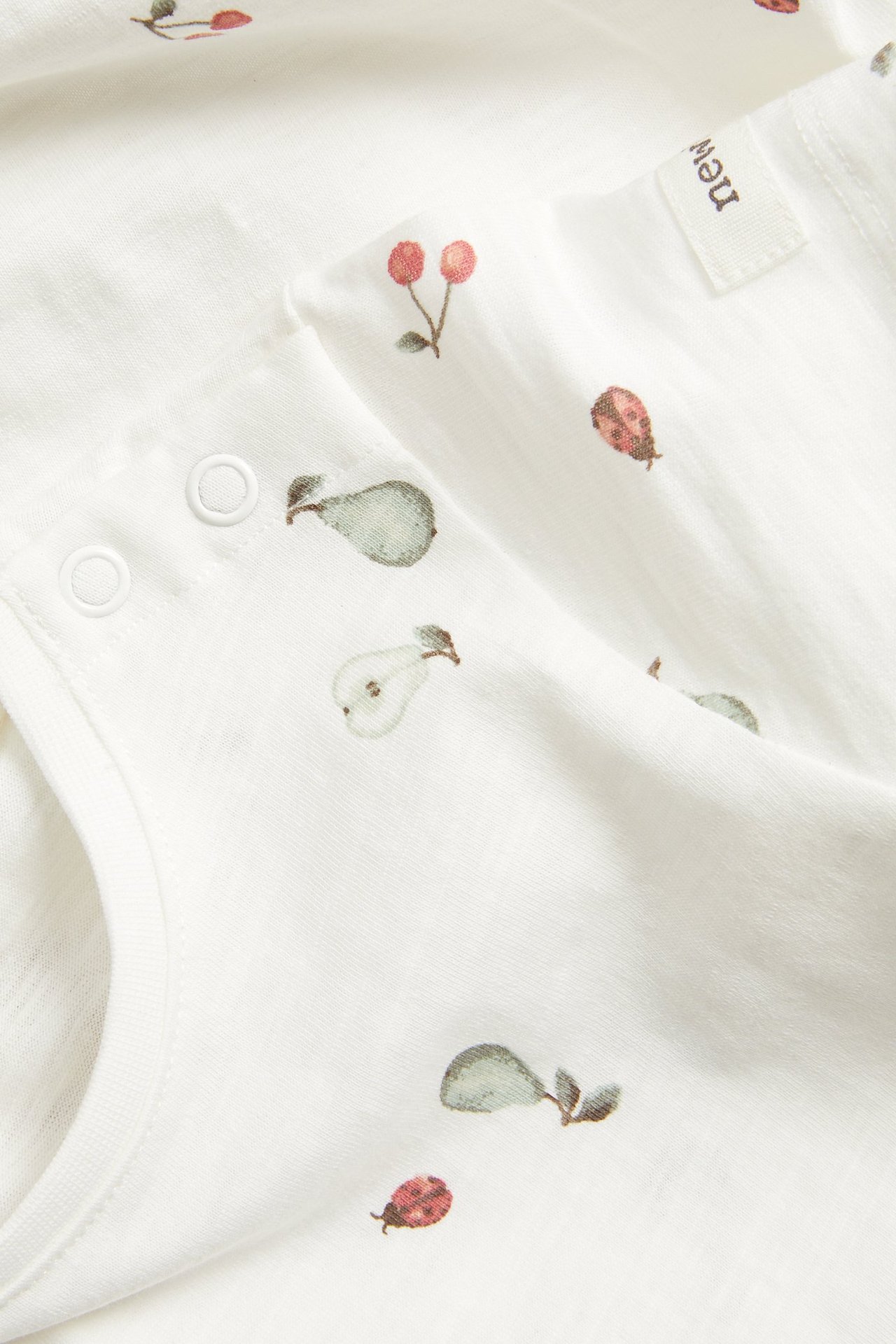 Nahaufnahme eines cremeweißen Baby-Bodys aus Baumwollslub mit Birnen-, Kirsch- und Marienkäferdruck, Druckknöpfen.