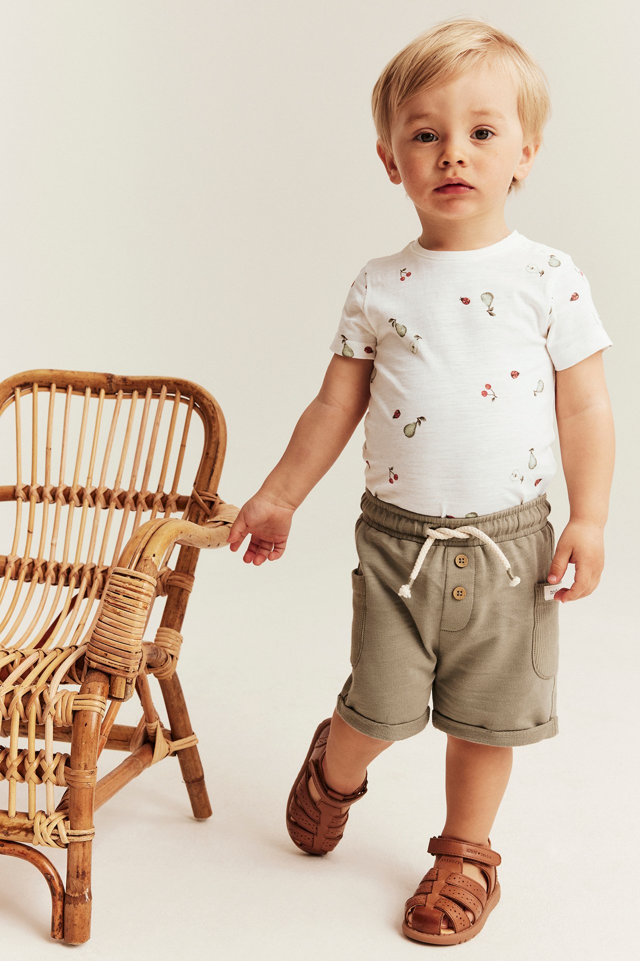 Front view: baby in off-white cotton short-sleeved bodysuit with fruit pattern, olive shorts, brown sandals.