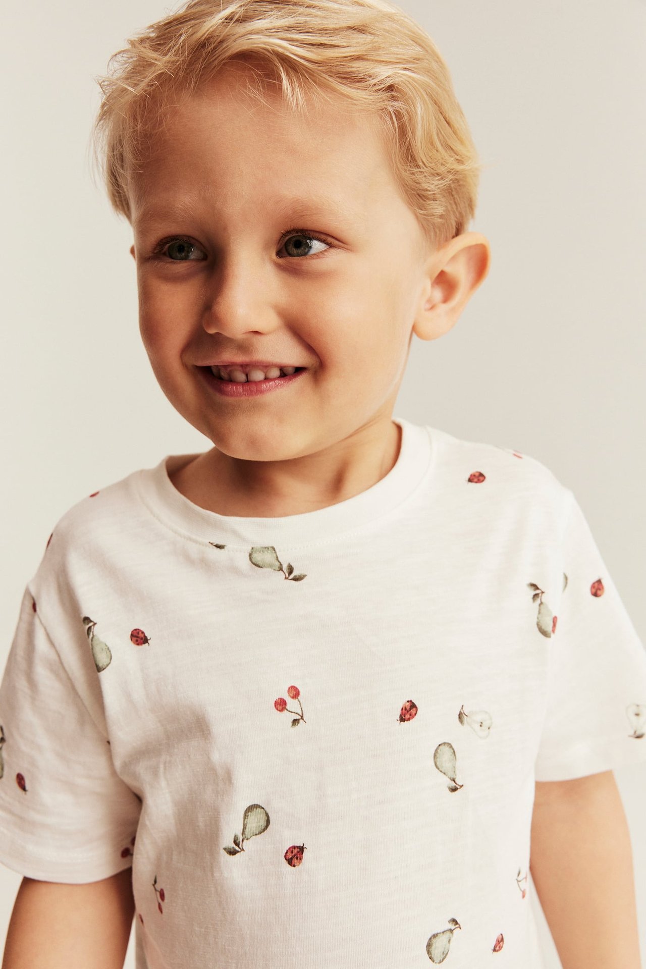 Vorderansicht eines lächelnden Jungen in einem cremefarbenen Slub-Jersey-T-Shirt mit Birnen-, Kirsch- und Marienkäferdruck.