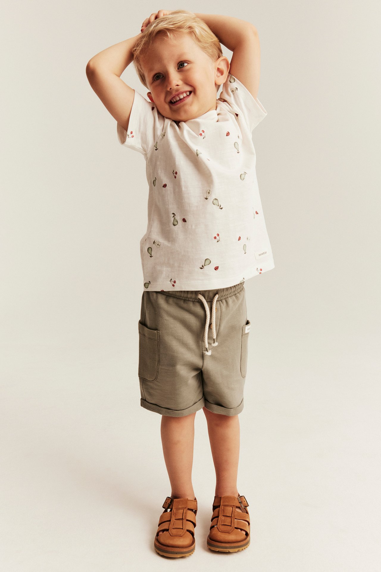 Front view of a kid wearing an off-white fruit-patterned t-shirt, khaki shorts, and brown sandals.