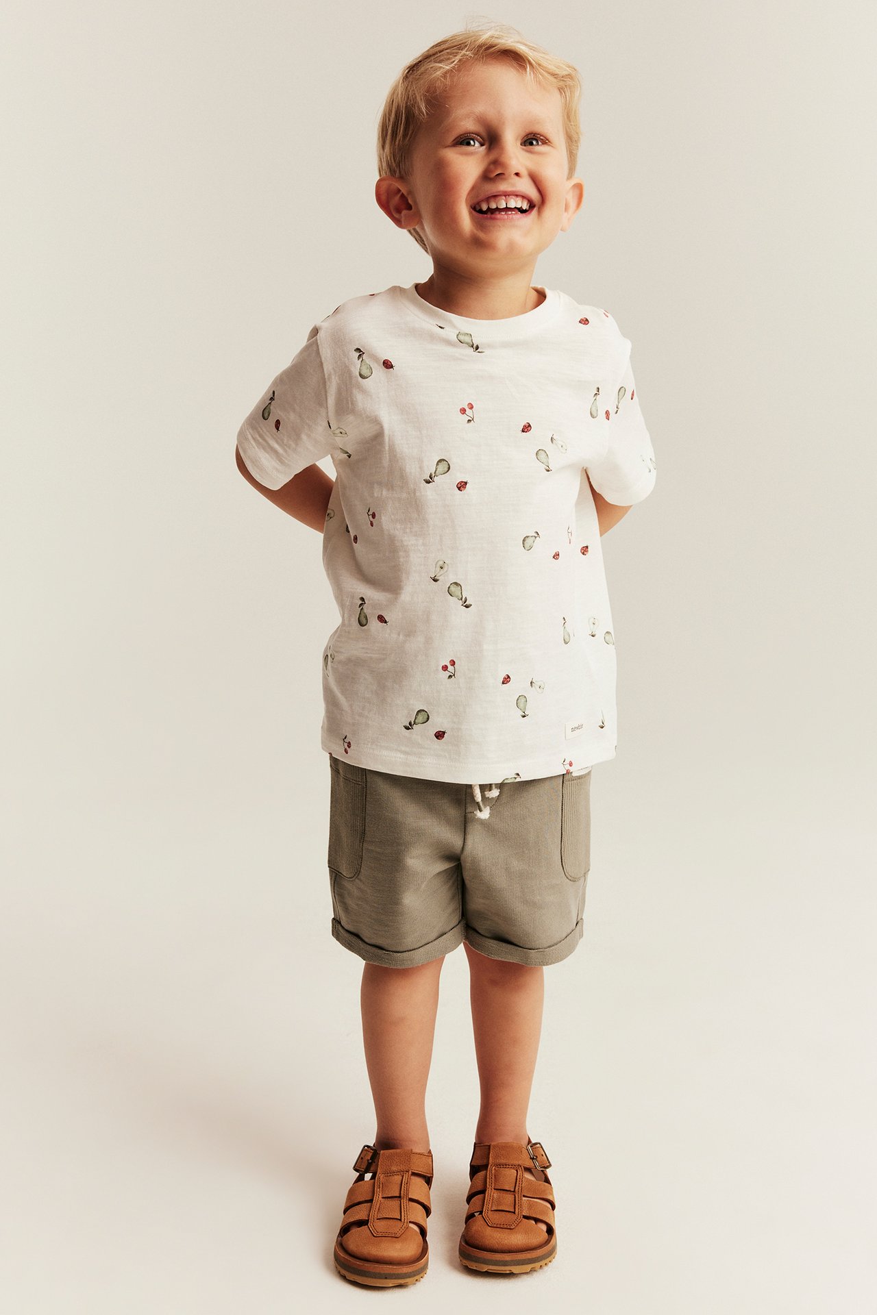 Smiling kid in offwhite t-shirt with pears, cherries, ladybugs, khaki shorts, and brown sandals.