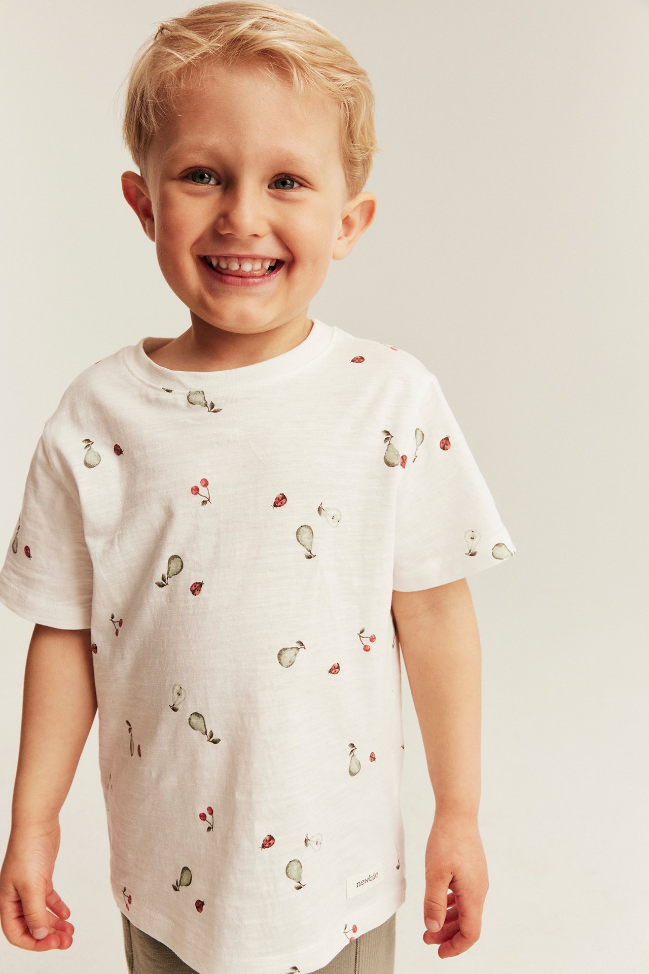 Front view: Smiling kid in an off-white t-shirt patterned with pears, cherries, and ladybugs, wearing grey pants.