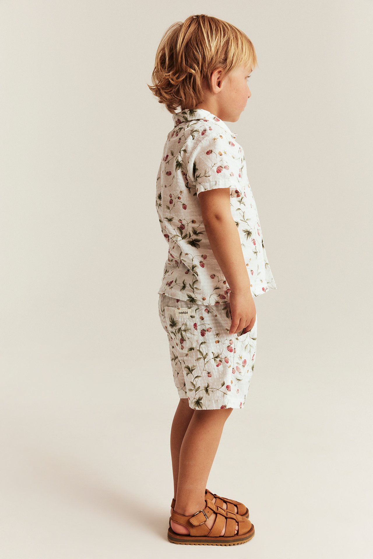Seitenansicht eines Kindes in offwhite gewebten Baumwollshorts mit Erdbeermuster, passendem Hemd und braunen Sandalen.