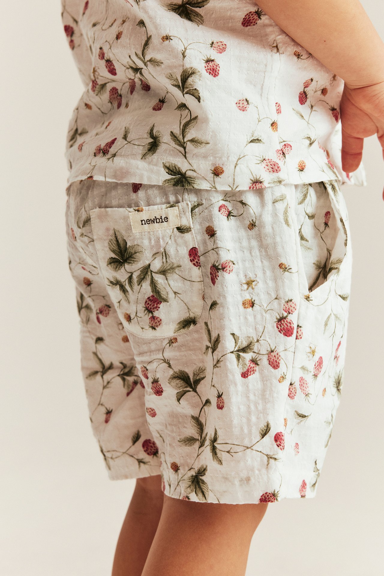 Seitenansicht eines Kindes: off-white gewebte Shorts mit Erdbeermuster und Taschen, dazu passendes Oberteil.