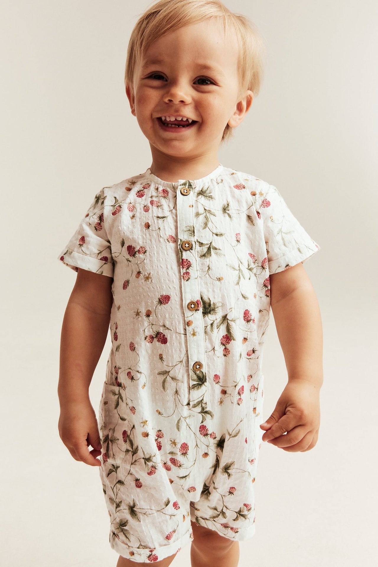 Frontansicht fröhliches Baby in offwhite gewebtem Baumwoll-Strampler mit Erdbeermuster, Knöpfen.