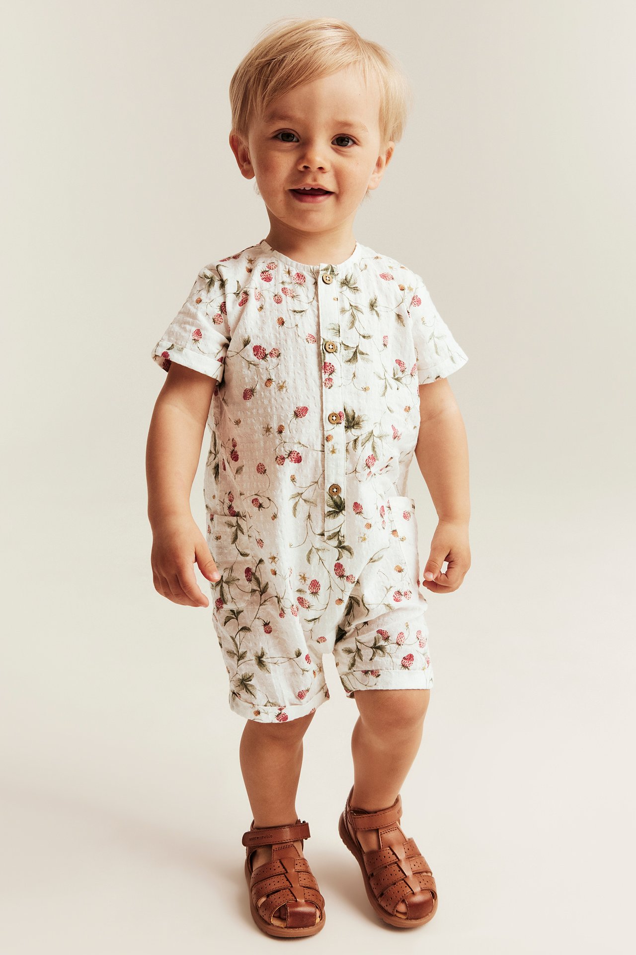 Vorderansicht, ein Baby trägt einen offwhite Erdbeermuster-Baumwollstrampler und braune Sandalen.
