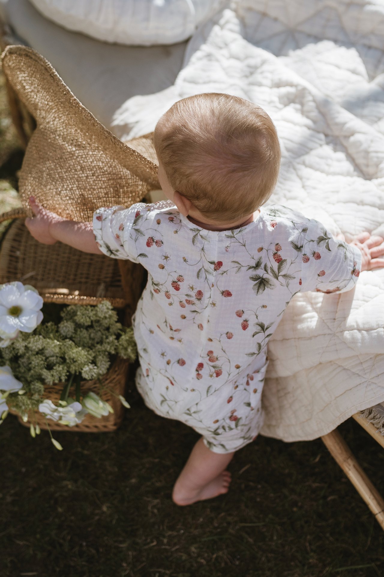 Von oben, hinten: Baby in cremeweißem Erdbeer-Baumwoll-Strampler, greift nach Strohhut.