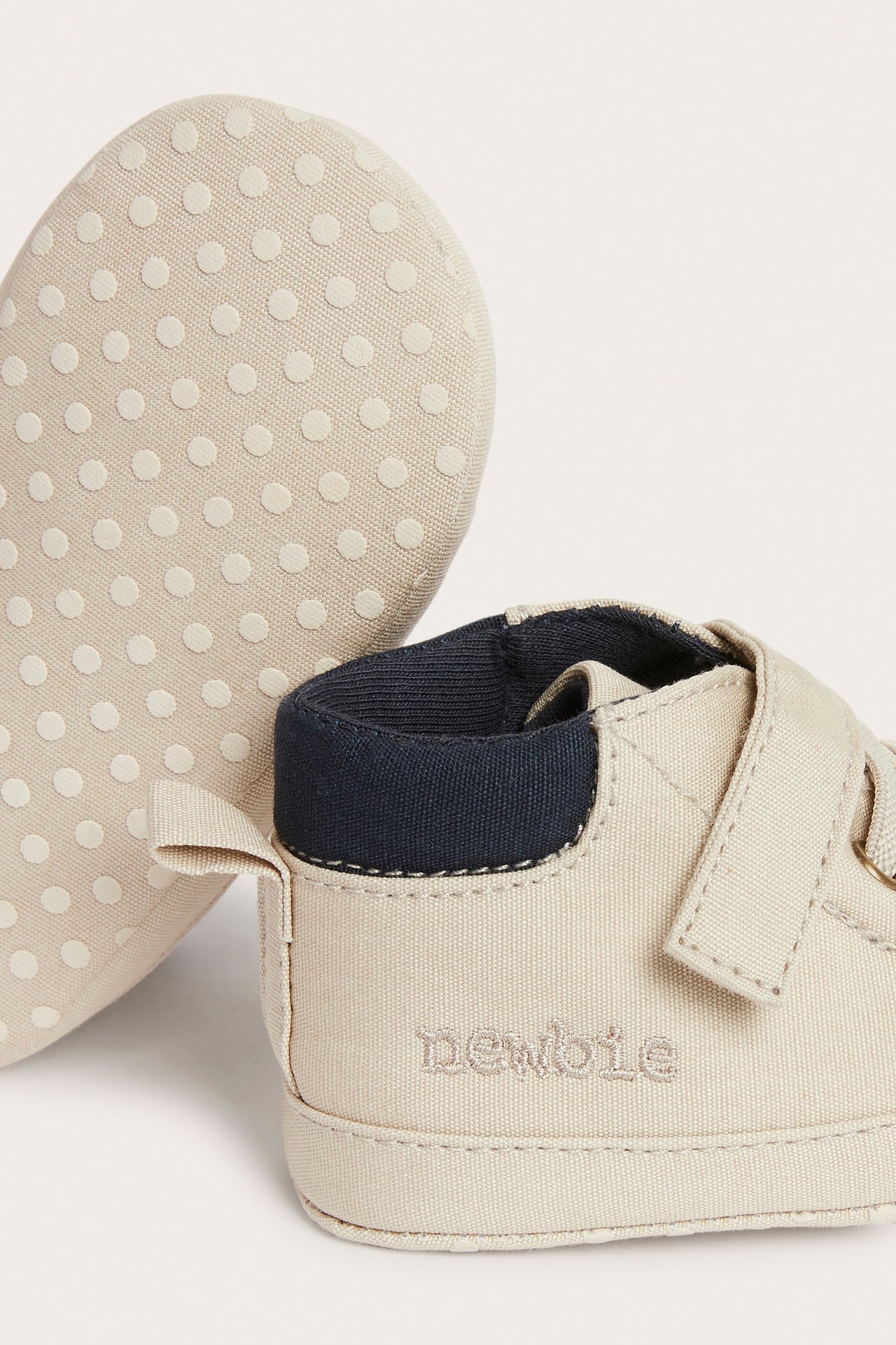 Nahaufnahme beiger Chambray-Babyschuhe mit Klettverschluss, elastischen Schnürsenkeln, Newbie-Stickerei und rutschfester Sohle.