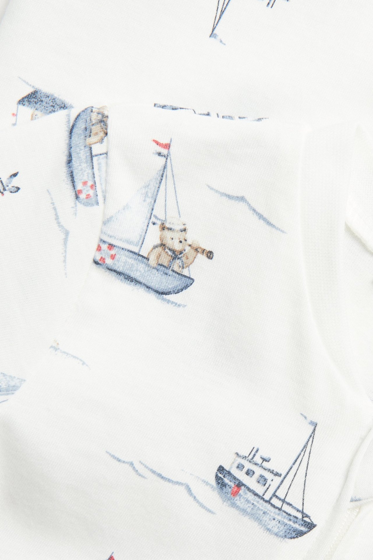 Close-up of an off-white cotton baby romper with a blue sailboat and teddy bear print.