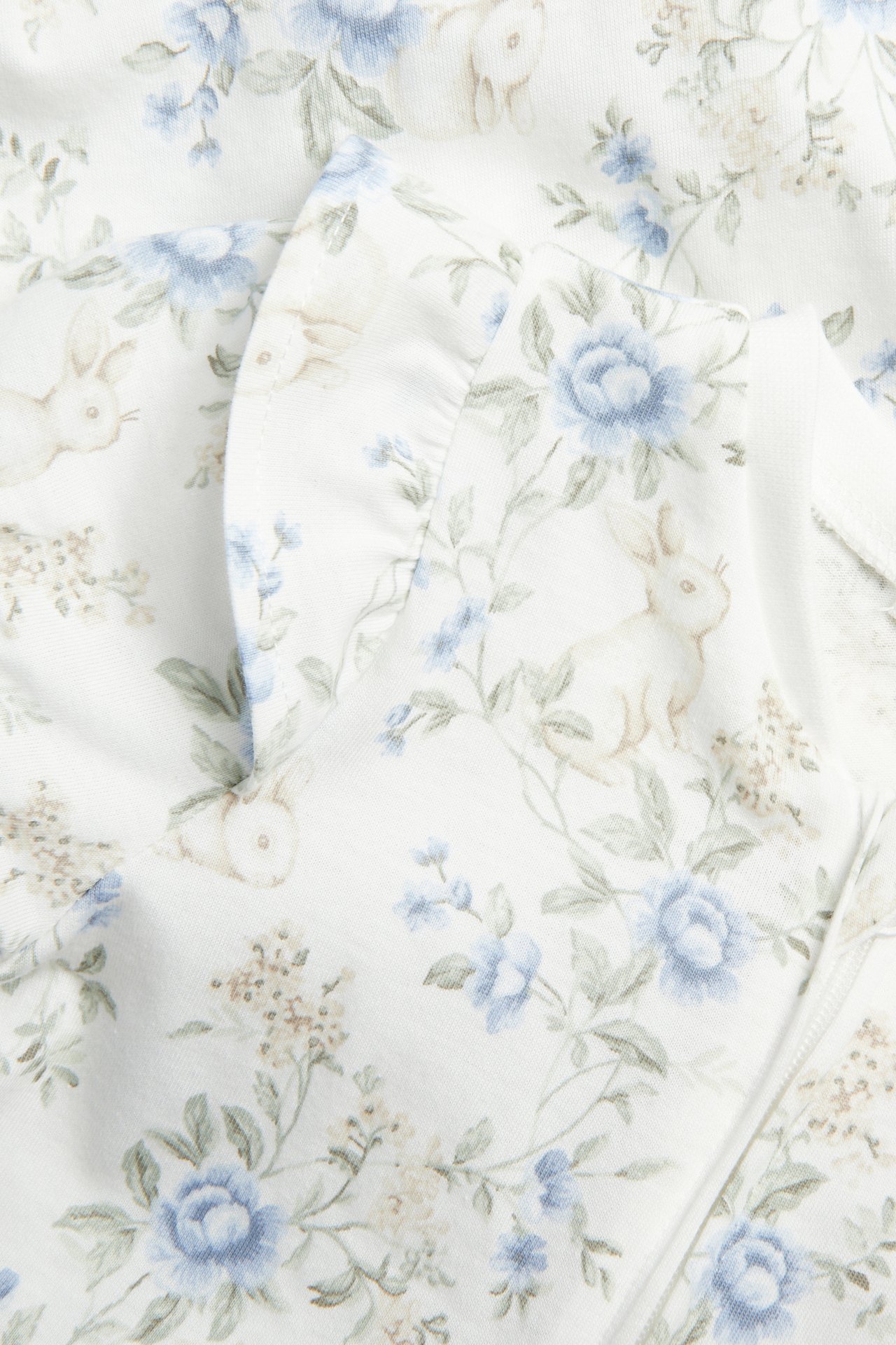 Close-up of an off-white baby pajama with light blue floral and bunny print, featuring a ruffled sleeve.