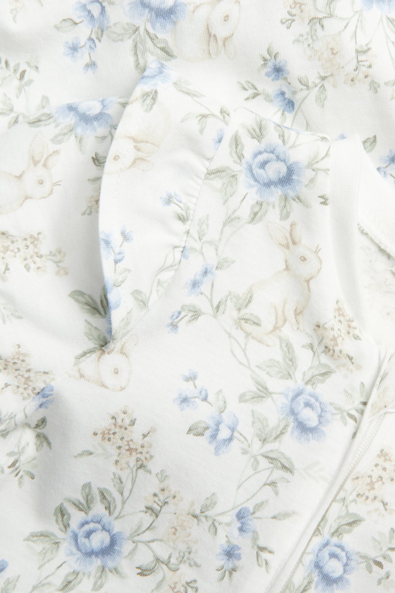Close-up of an off-white baby pajama with light blue floral and bunny print, featuring a ruffled sleeve.