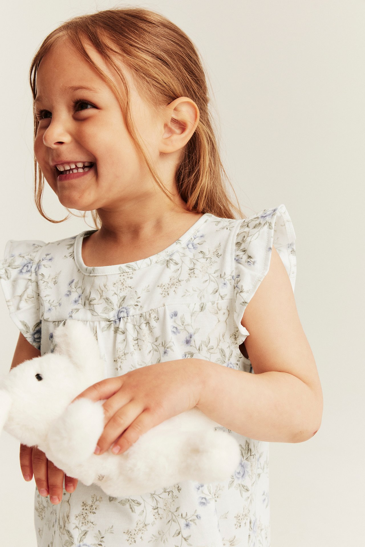 Front view of a kid in a white floral top with ruffled sleeves, holding a white plush bunny.