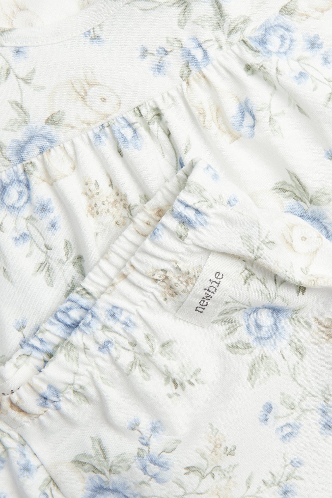 Nahaufnahme von cremeweißem Baumwollpyjama mit blauem Blumen- und Hasenmuster, zeigt geraffte Details.