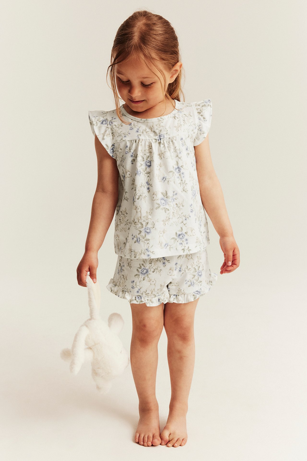 Front view of a kid wearing a light floral print short pajama set, holding a white plush bunny.