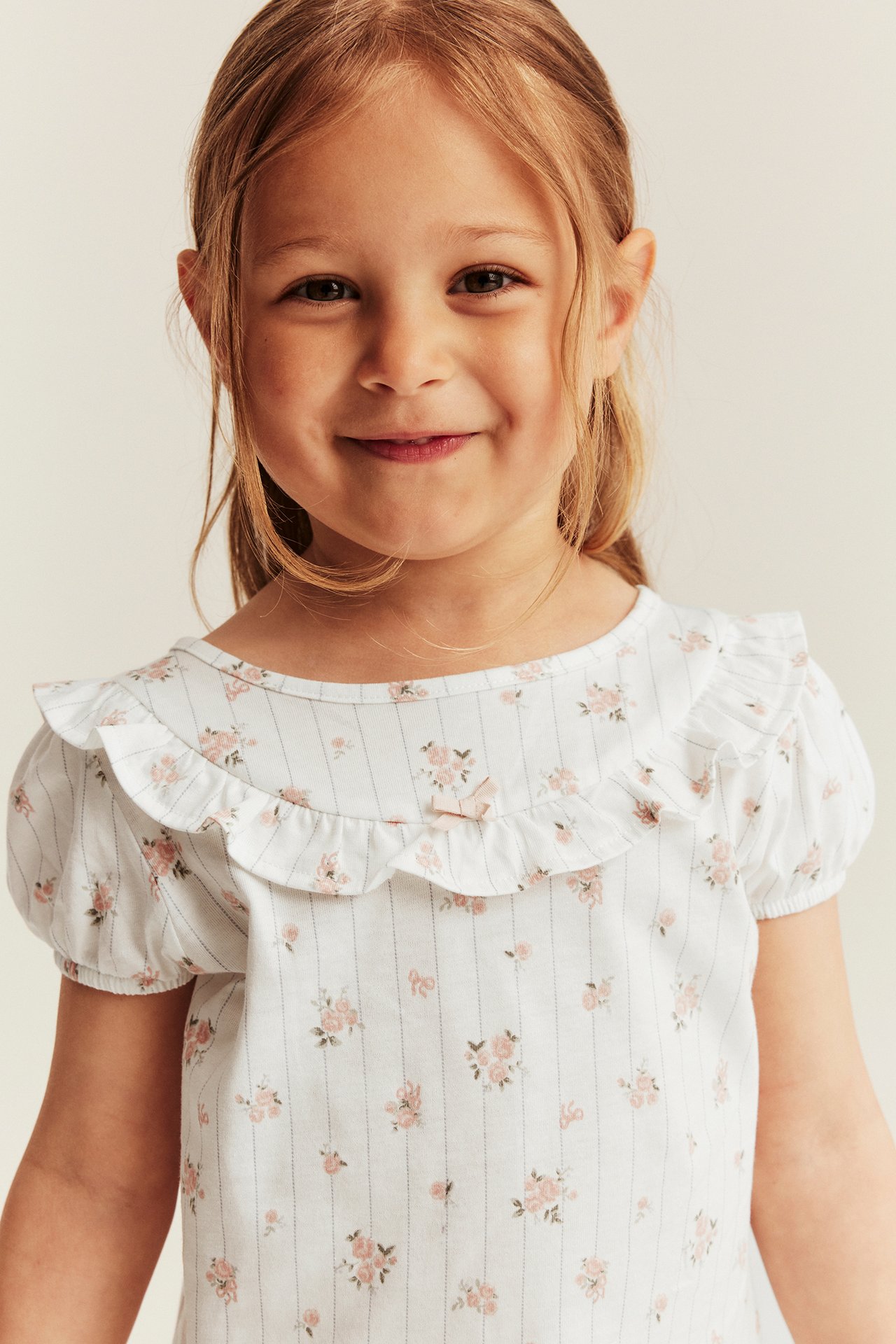 Front view: Kid wears off-white floral striped cotton pajama top with ruffles and a bow.