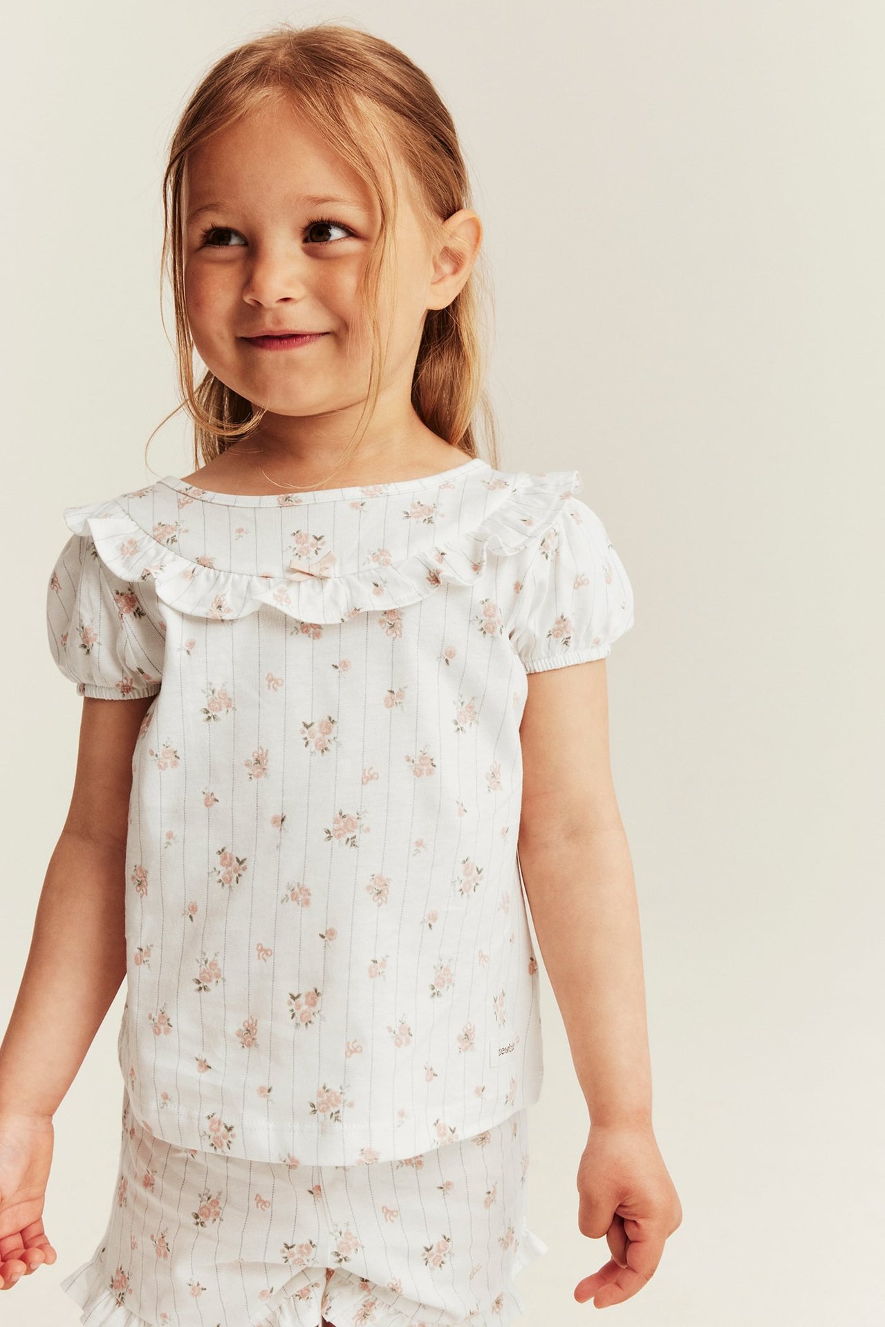 Vorderansicht: Kind trägt off-weißes, geblümtes und gestreiftes Baumwoll-Pyjama-Oberteil mit Rüschen und passende Shorts.