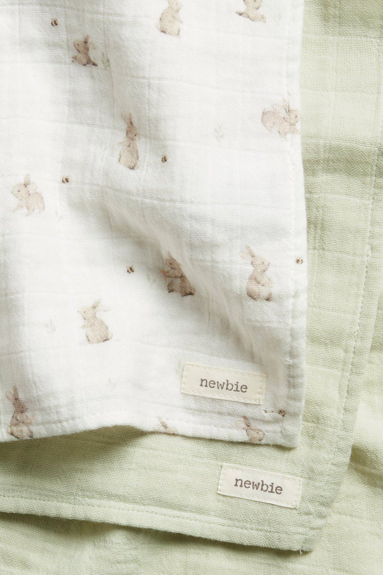 Close-up of two soft cotton muslin blankets: one off-white with rabbit print, one solid light green.