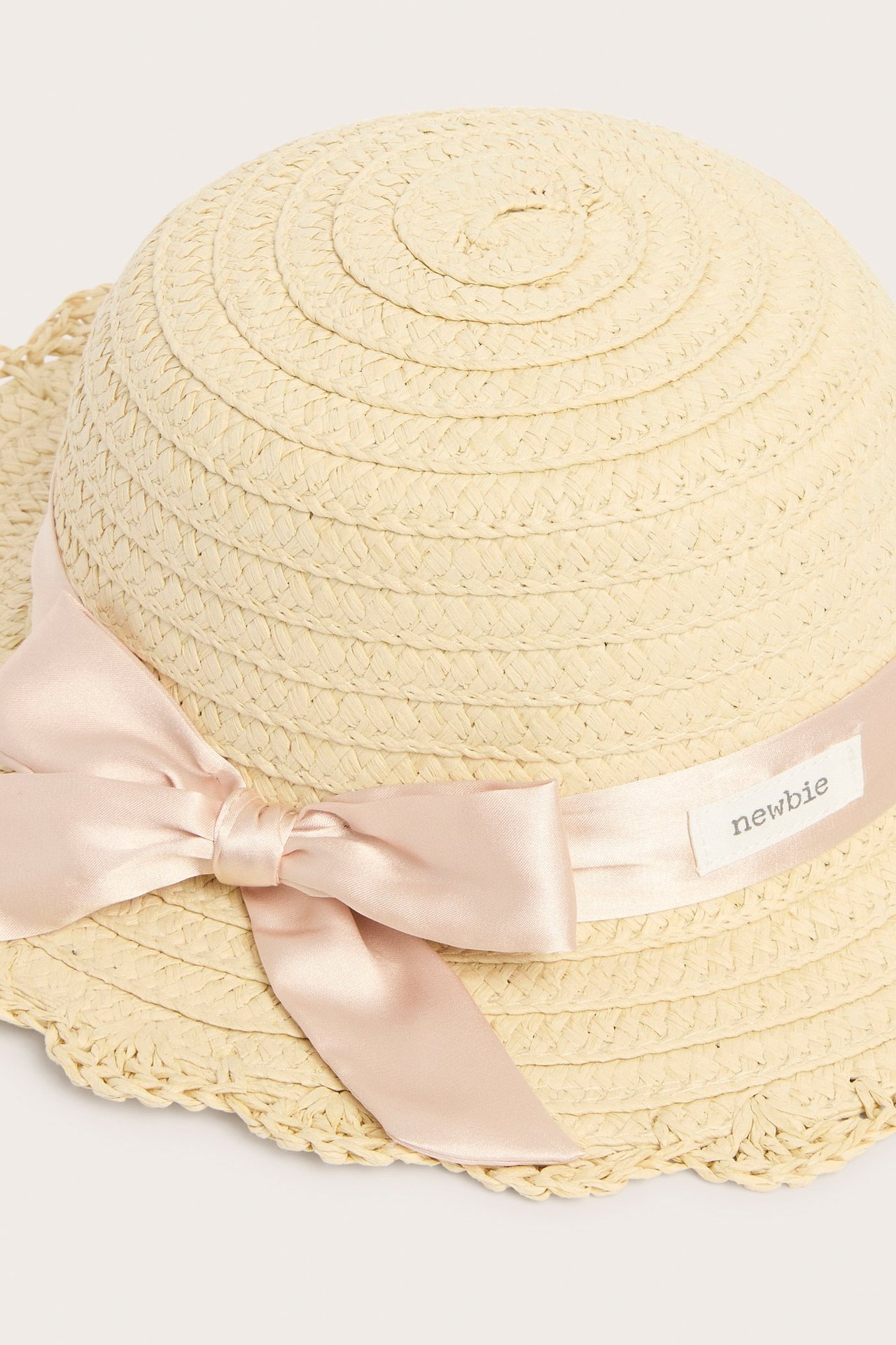 Close-up of a light beige woven straw hat with a pink satin bow and 'Newbie' label.