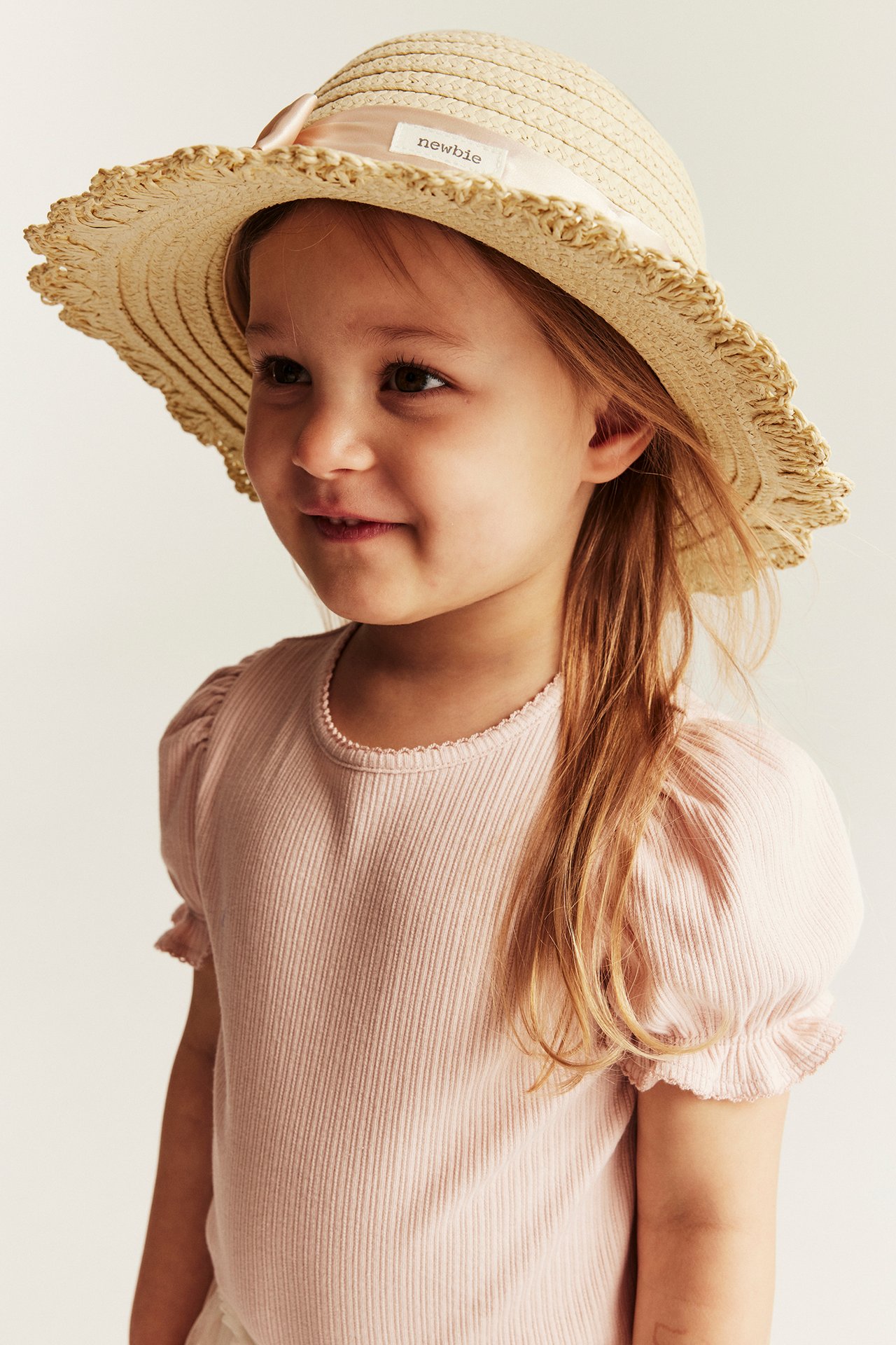 Side view: kid in light beige structured straw hat with pink bow, and a pink ribbed short-sleeve top.