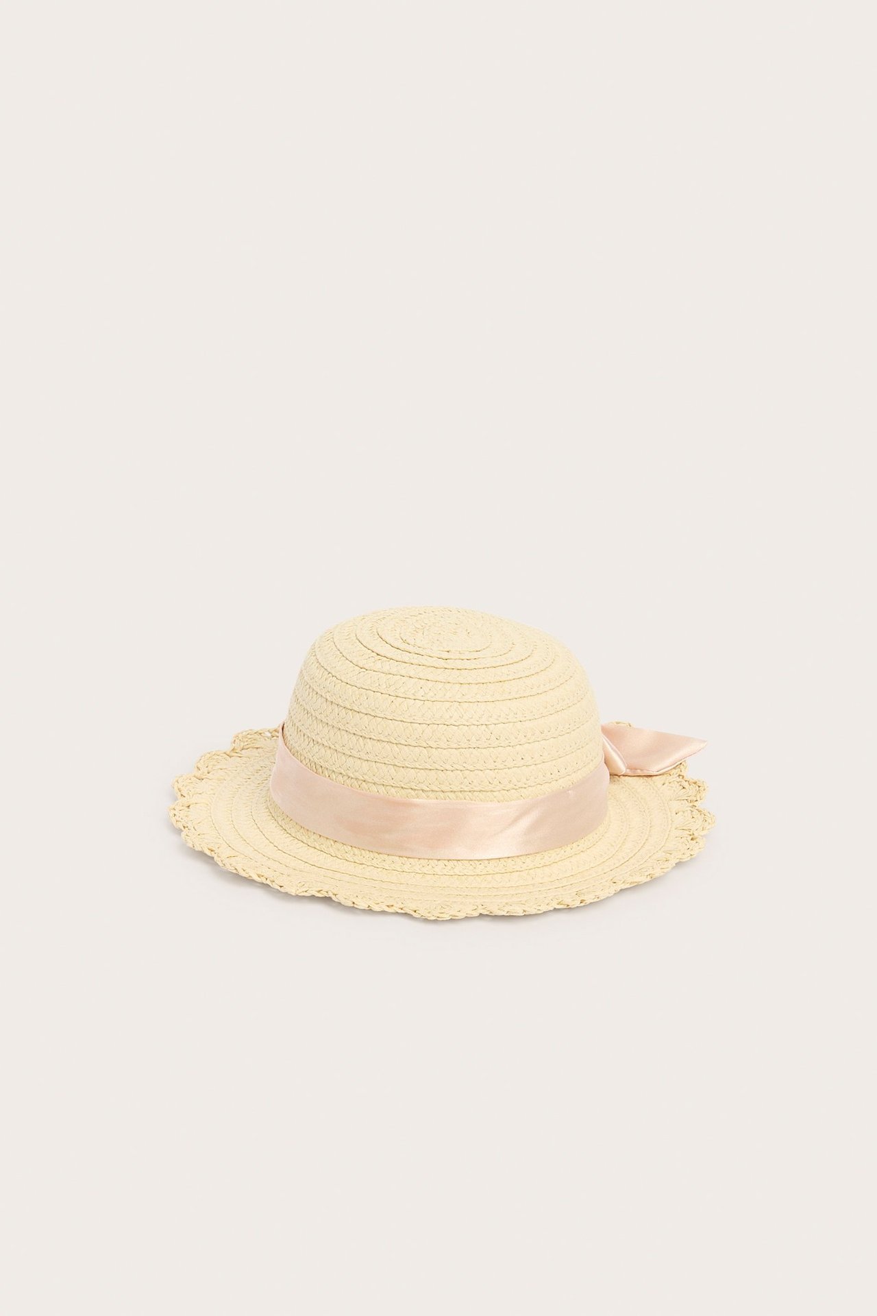 Front-side view of a light beige straw hat for kids with a light pink satin bow and scalloped brim.